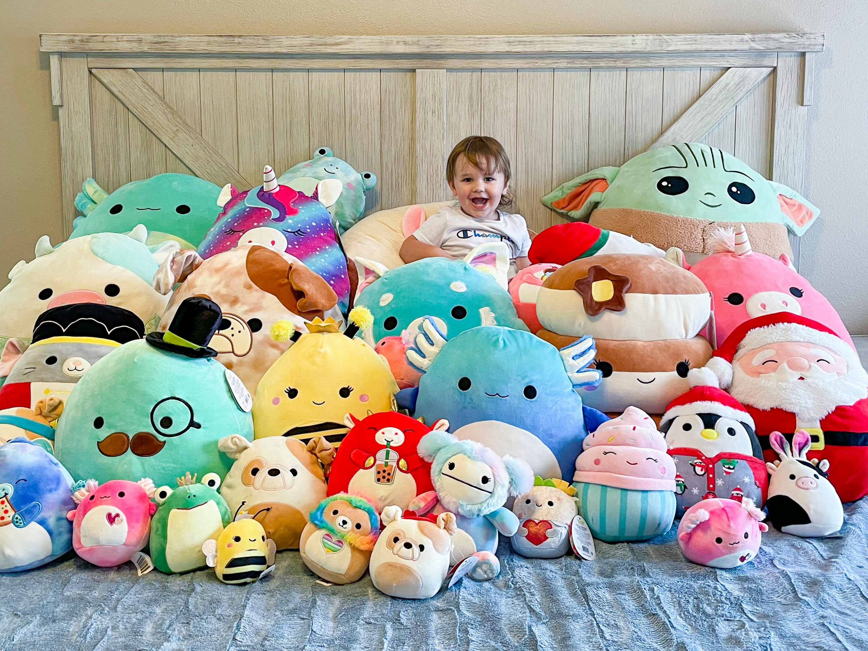 A child sitting on a bed covered in a huge pile of different sized Squishmallows.