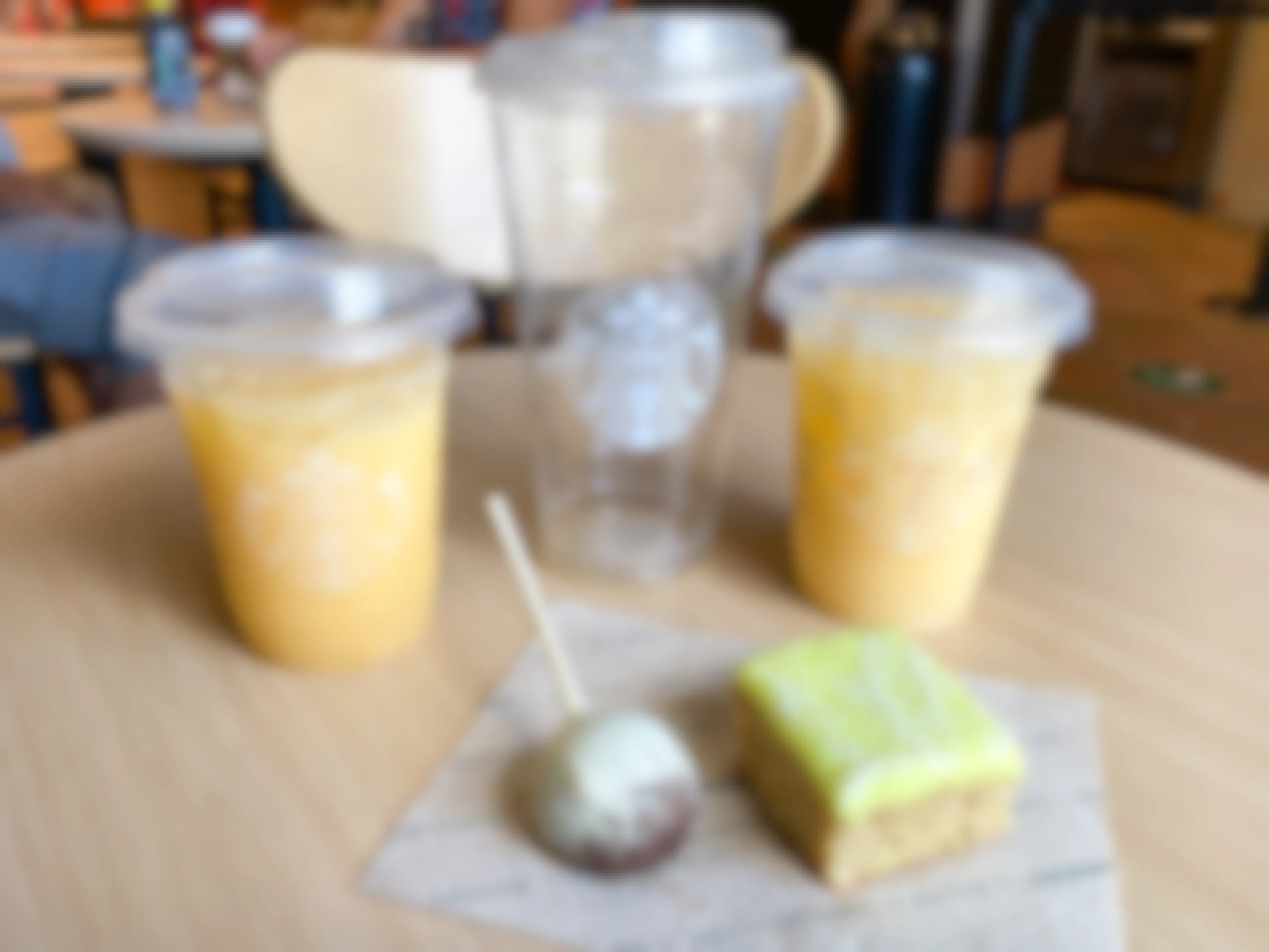 Two tall Starbucks Paradise drinks and an empty venti Starbucks cup sitting next to a cookies and cream cake pop and a lime-frosted cake square on a table in Starbucks.