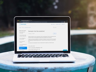 A laptop sitting on a table by a pool, displaying the Host Fee explanation page on the Swimply wesbite.