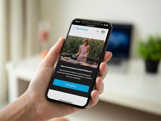 A person's hand holding a cell phone displaying the Swimply spaces page on their app.