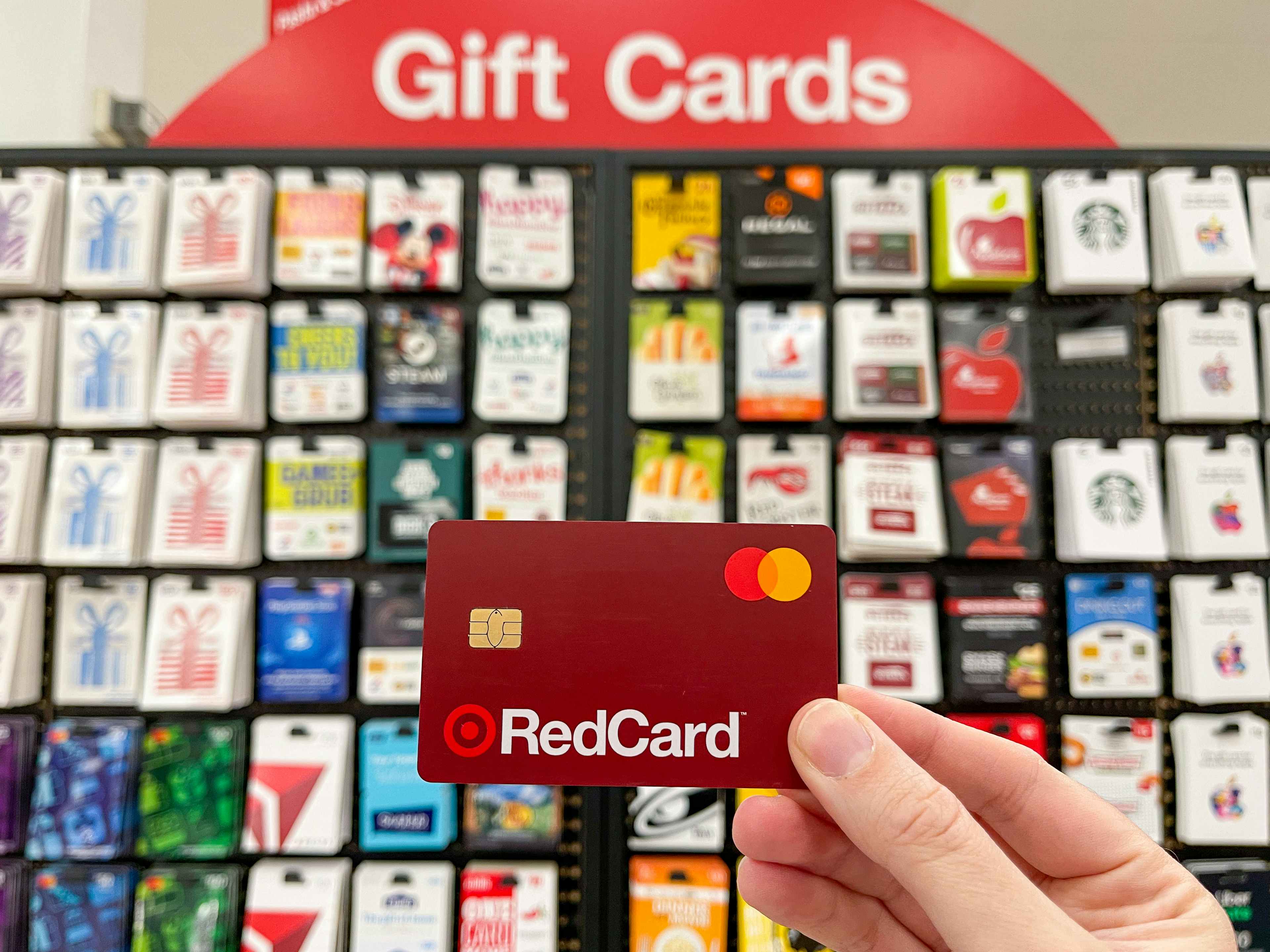 A person's hand holding their RedCard in front of wall display of gift cards for stores, restaurants, and more inside a Target.