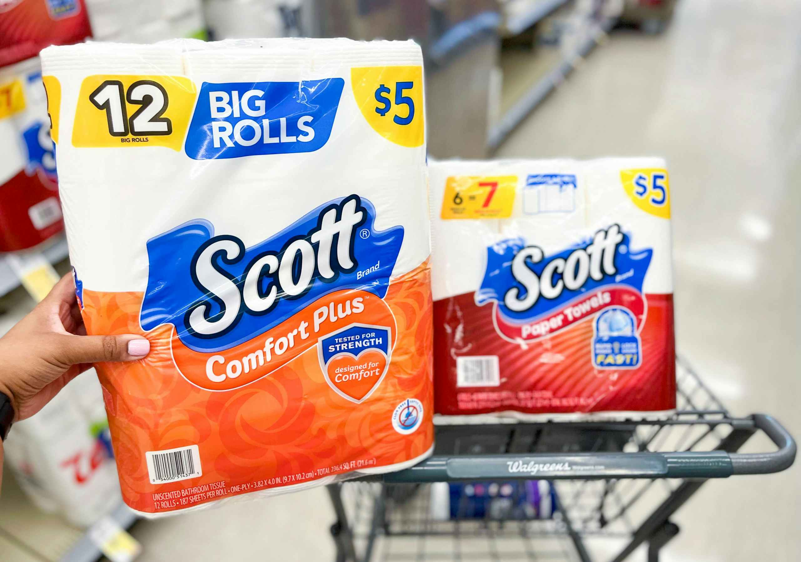 Hand holding a pack of Scott toilet paper with cart in the back with a pack of Scott Paper Towels inside of it