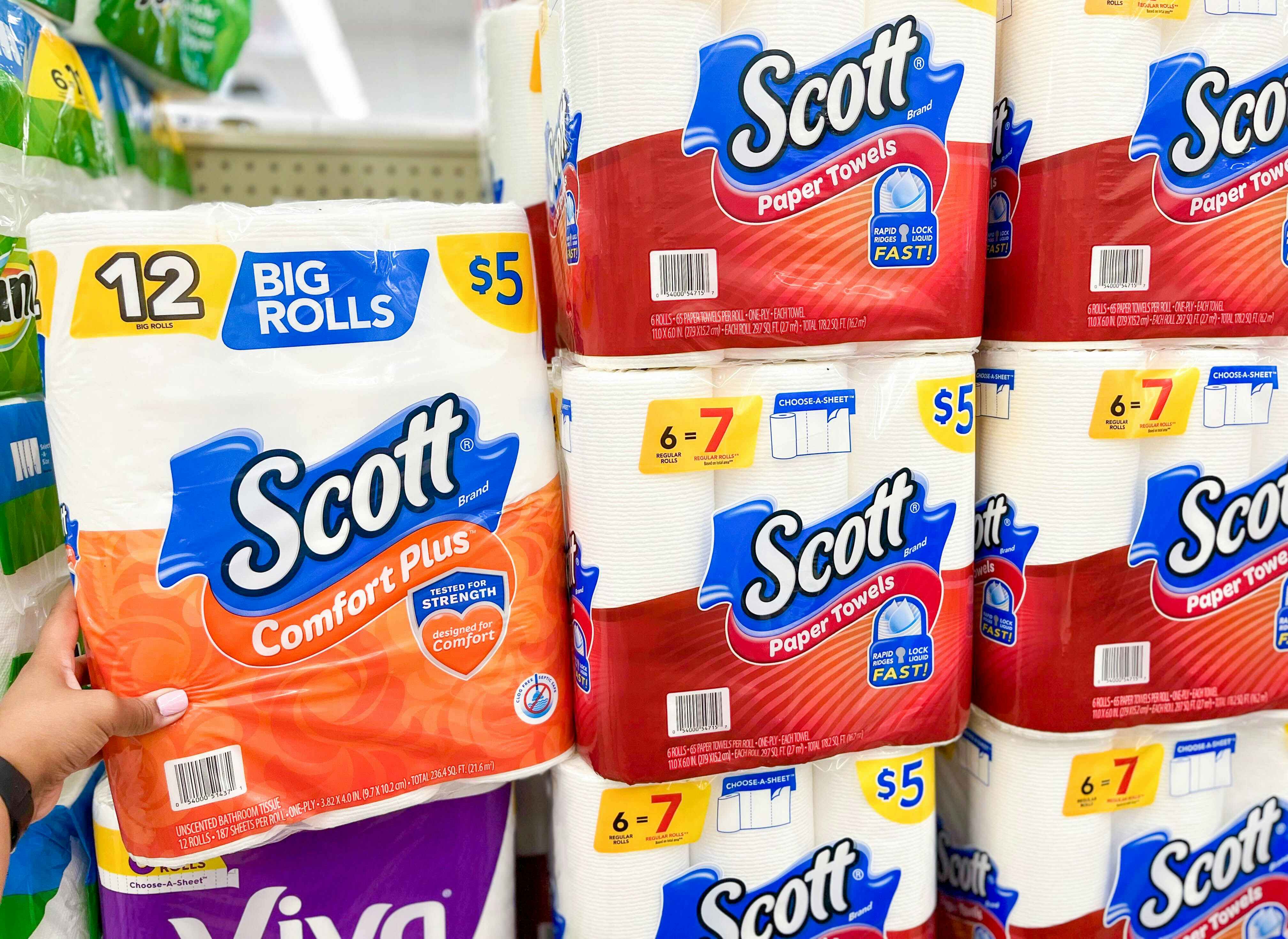 Hand holding a pack of Scott Toilet Paper next to a stack of Scott Paper Towel Packs