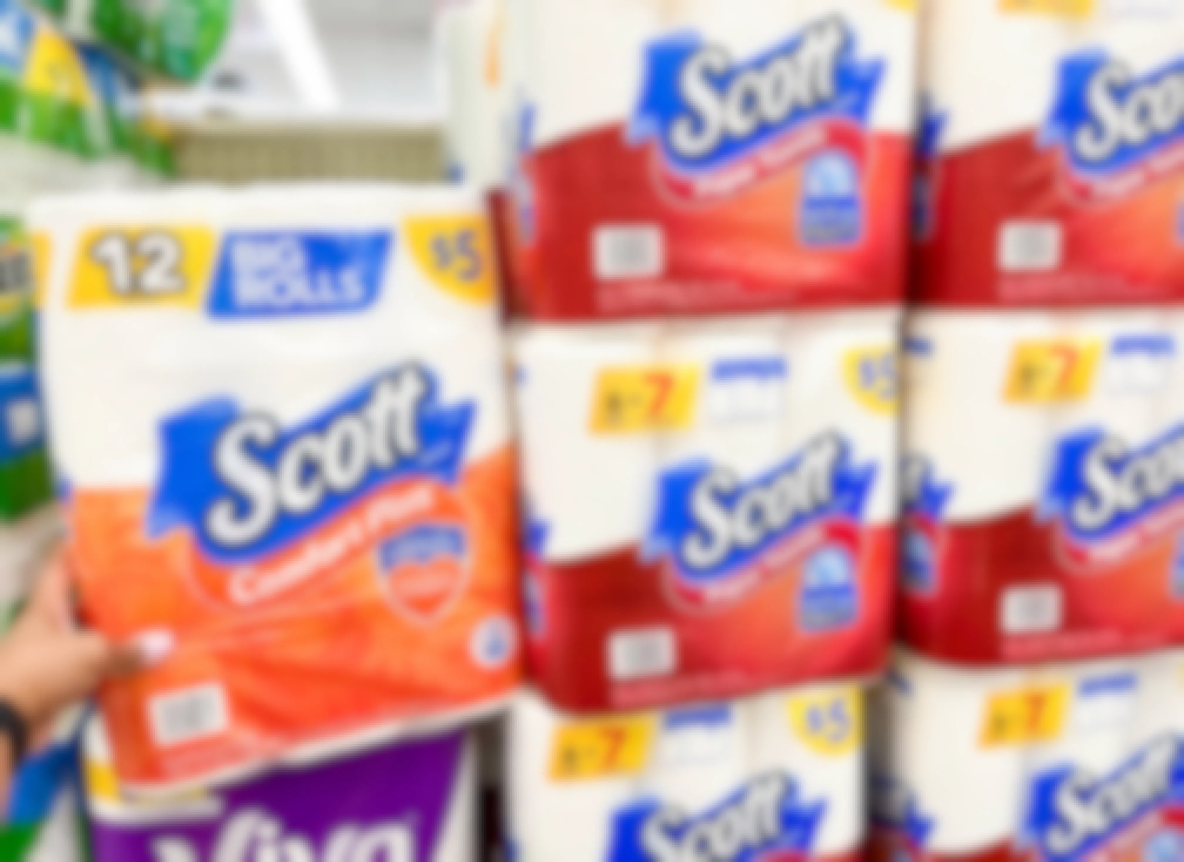 Hand holding a pack of Scott Toilet Paper next to a stack of Scott Paper Towel Packs