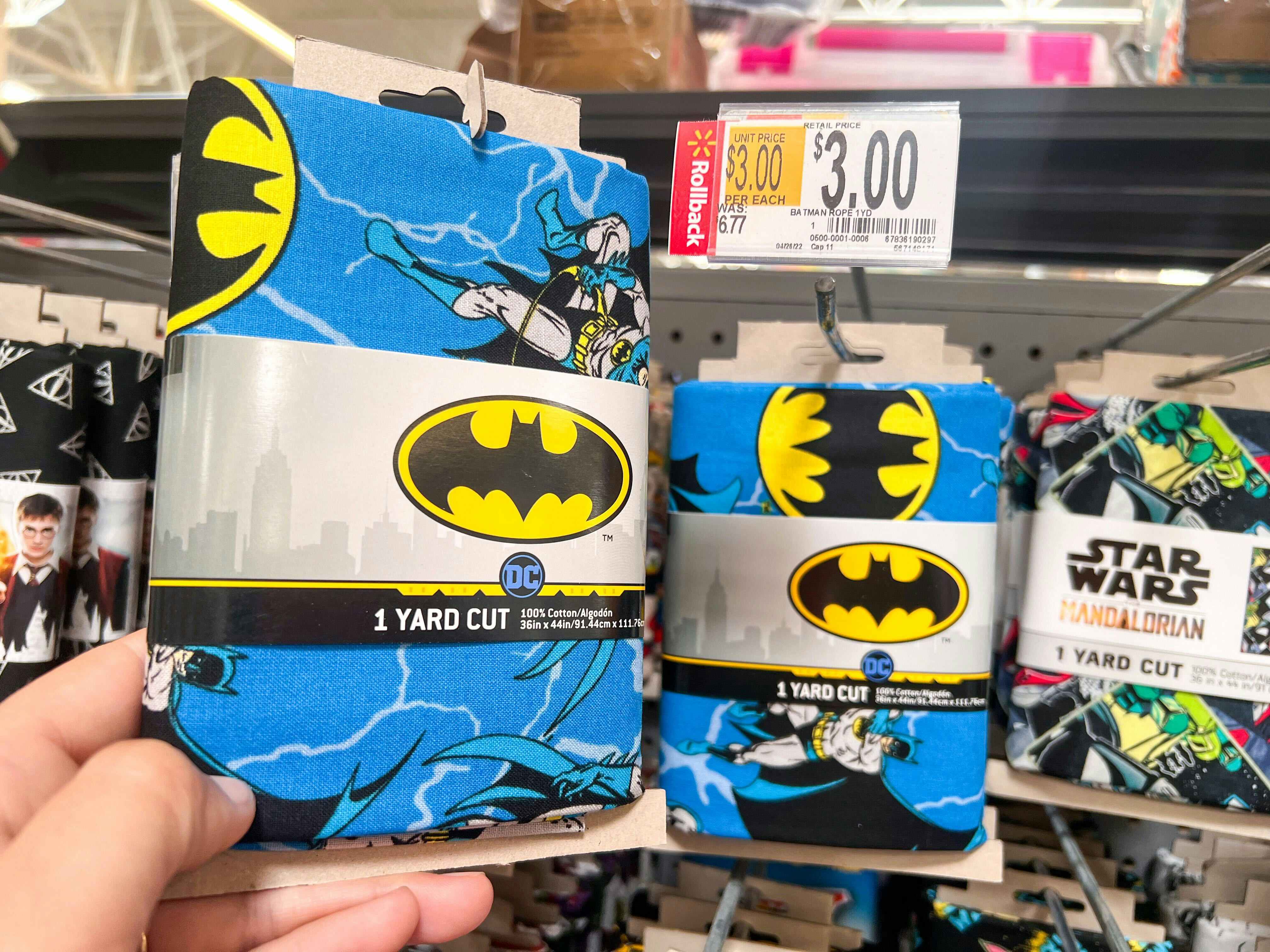 Hand holding Batman printed quilting fabric in front of shelf at Walmart. Price tag indicates that the product is on sale for $3, regularly $6.77.