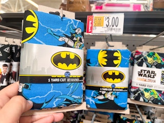 Hand holding Batman printed quilting fabric in front of shelf at Walmart. Price tag indicates that the product is on sale for $3, regularly $6.77.