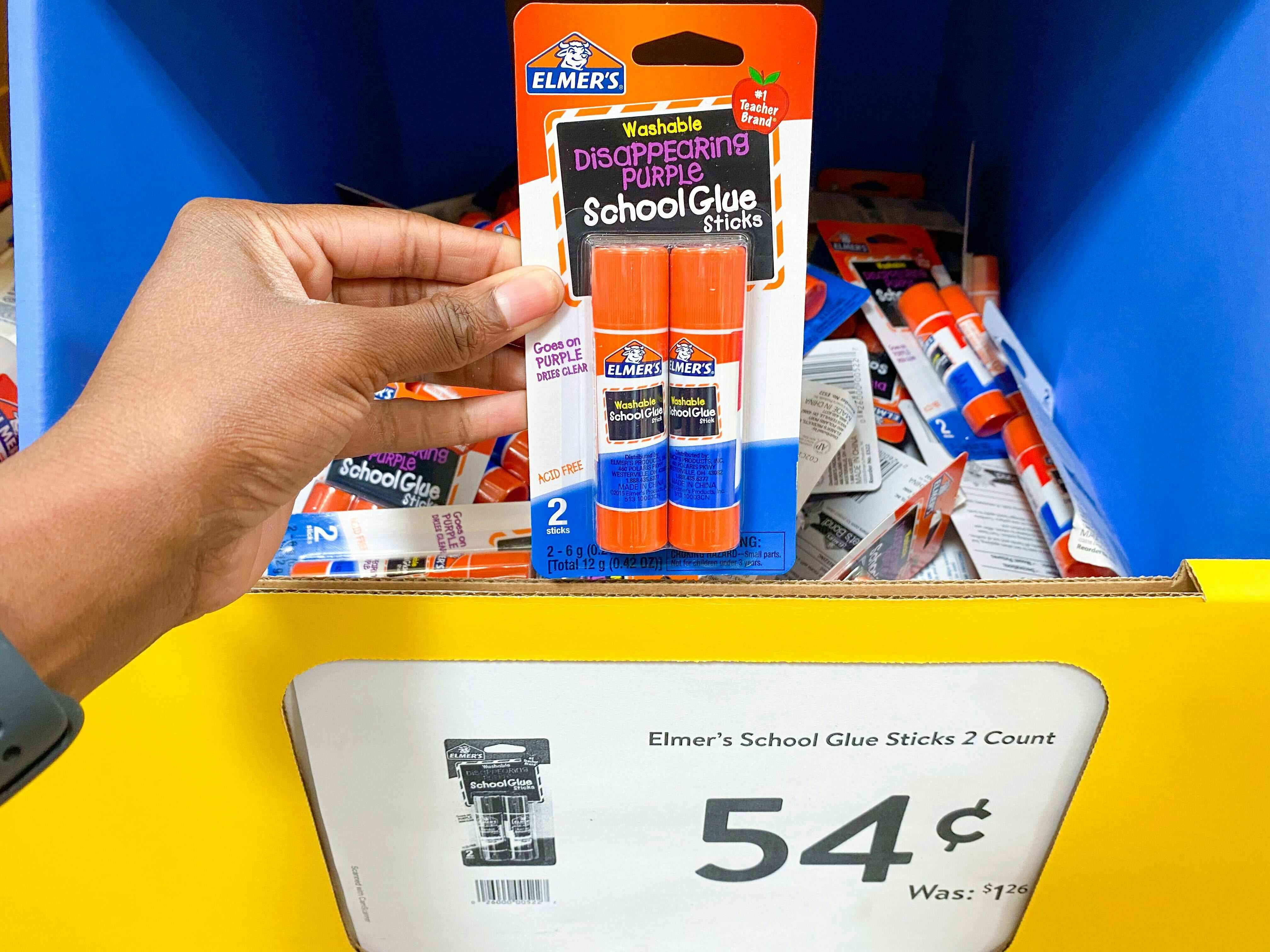 A person's hand holding a 2-pack of Elmer's glue sticks in front of a bin of more glue sticks with a sale tag pricing them at 54 cents.