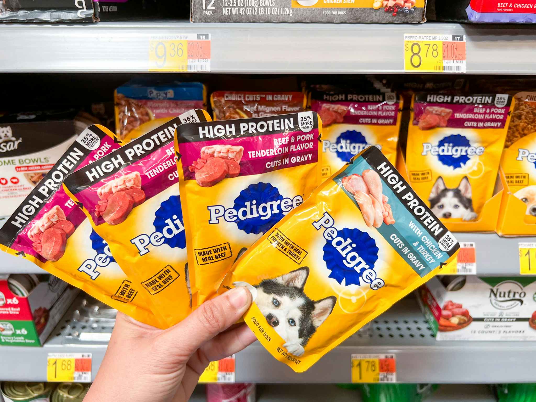 A hand holding four packs of pedigree high protein dog food in front of a walmart shelf