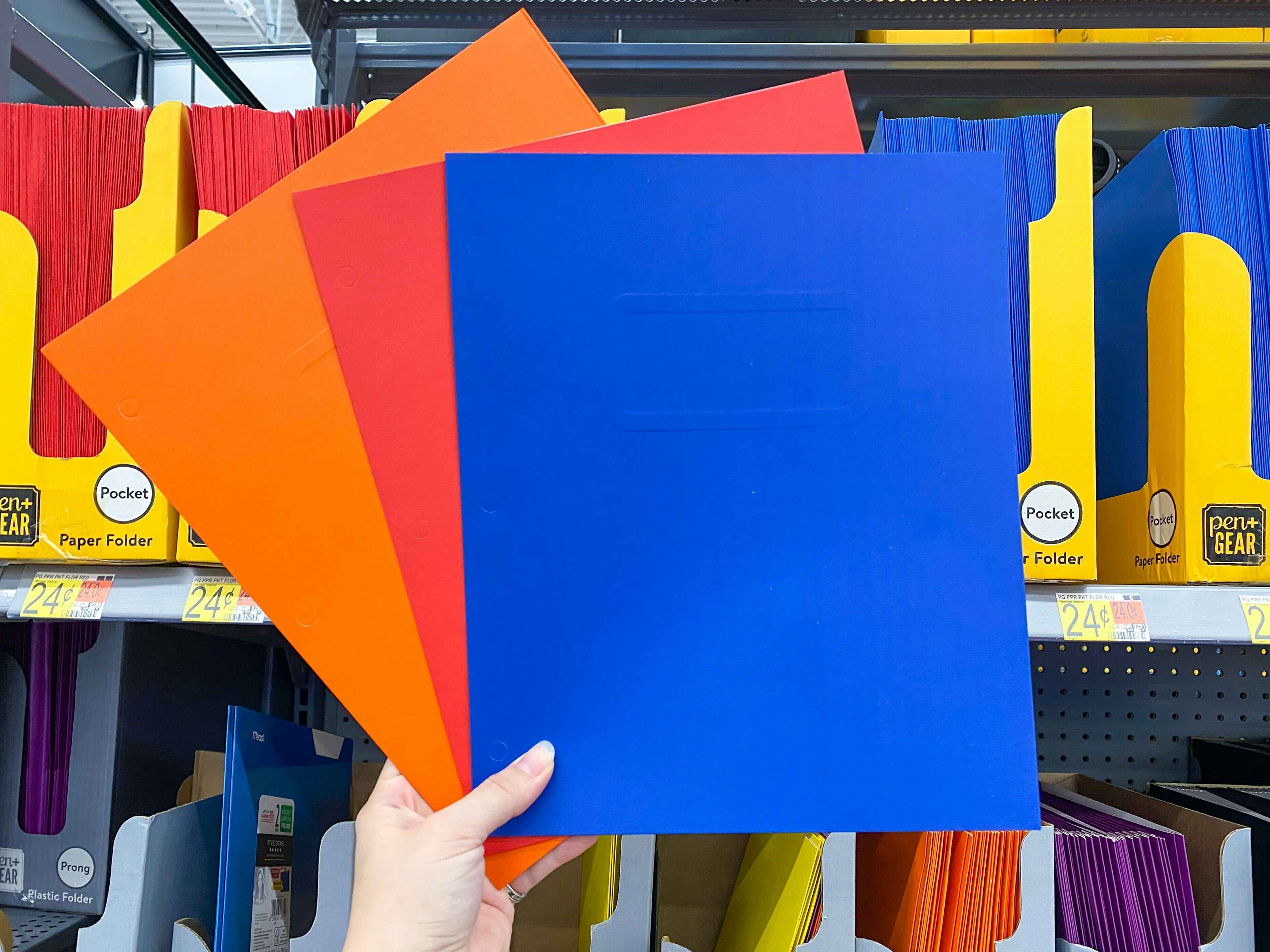 A person's hand holding up three different colored Pen+Gear paper folders in front of a shelf at Walmart.