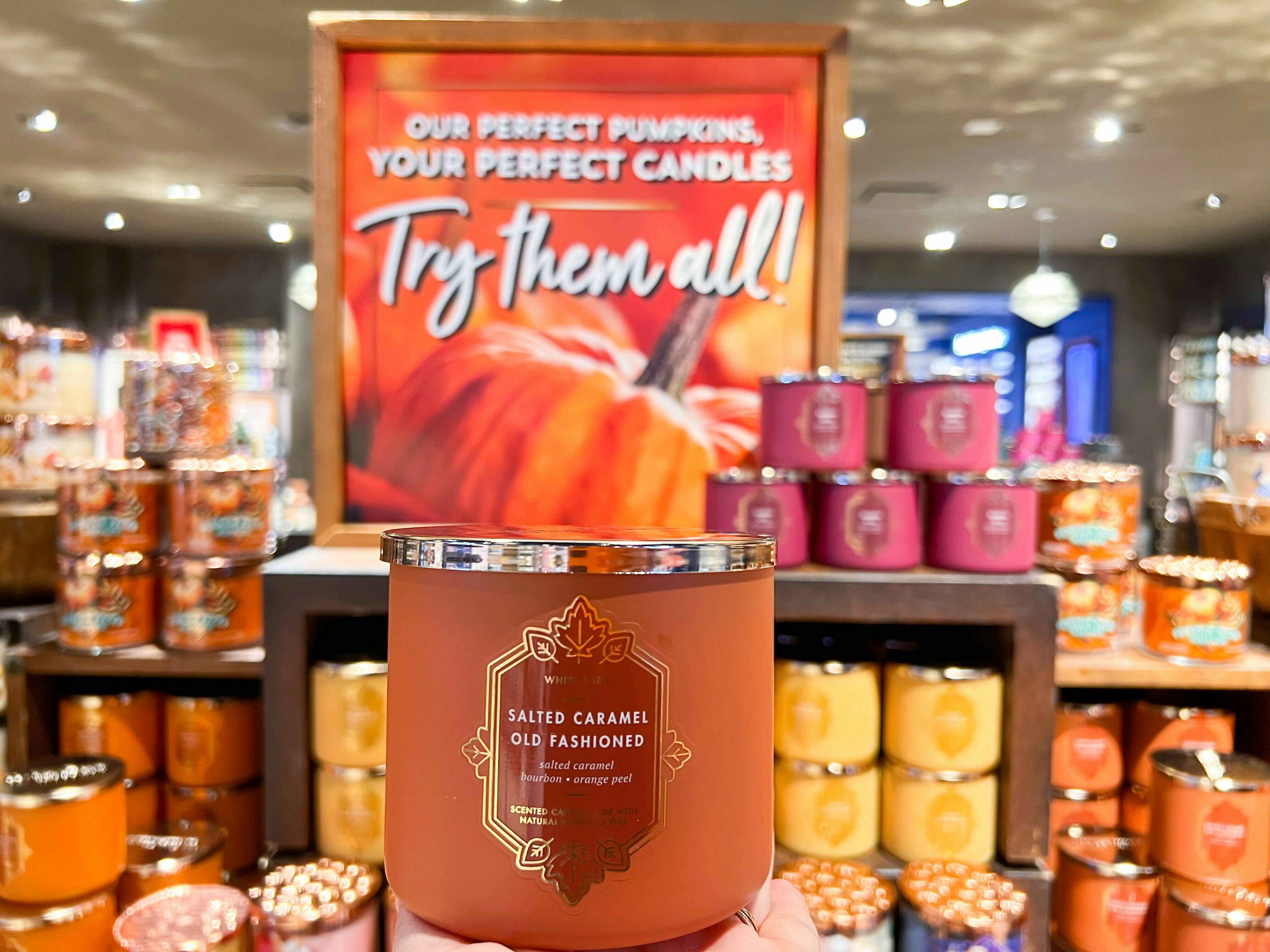 A person's hand holding up a fall 3-wick candle in front of a display of fall candles in Bath and Body Works.