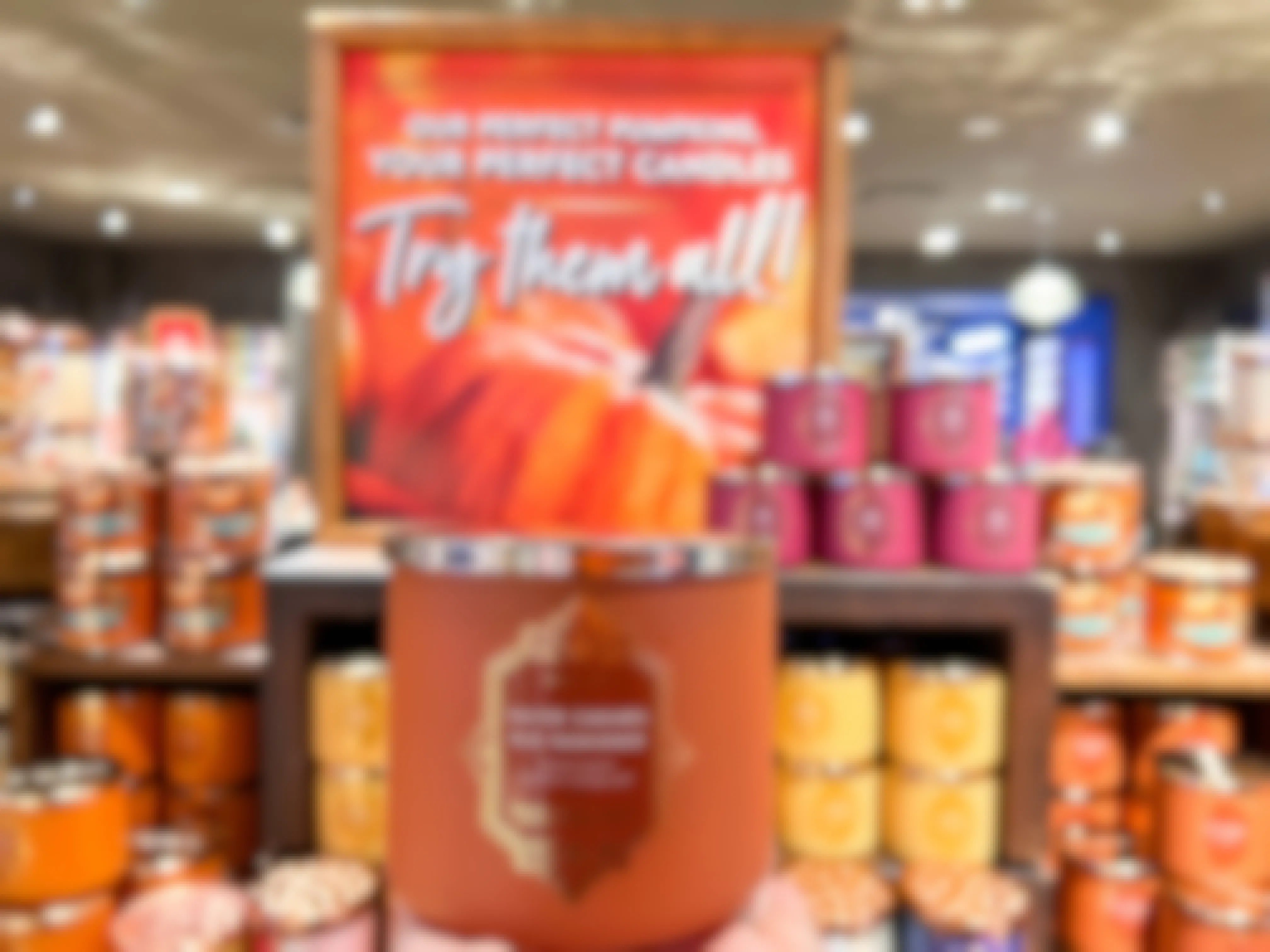 A person's hand holding up a fall 3-wick candle in front of a display of fall candles in Bath and Body Works.