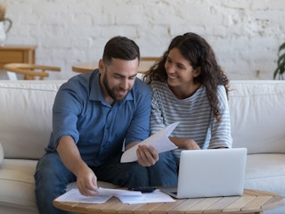 A couple sitting on a couch, smiling and holding bills with a laptop on the table next to them.