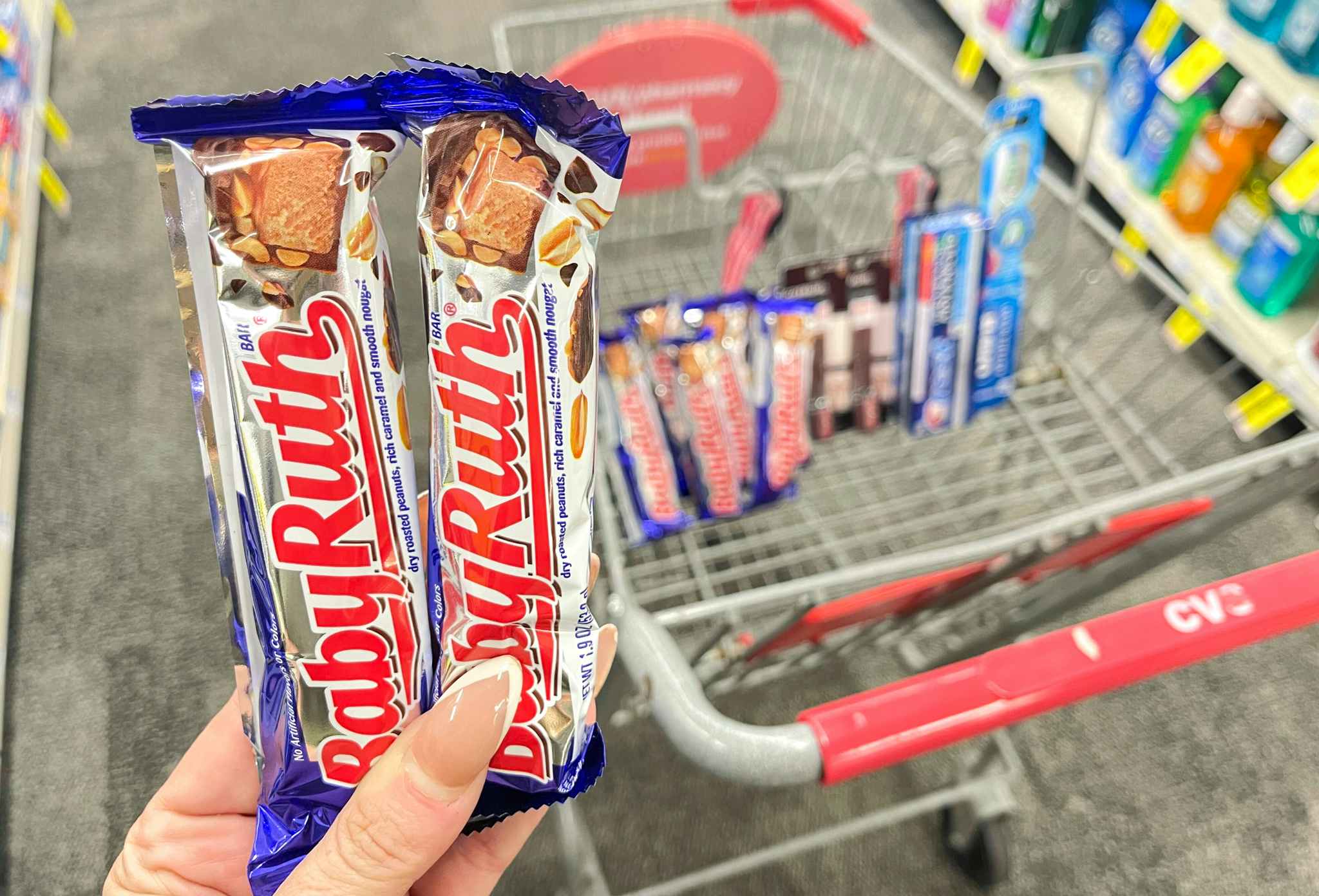 hand holding two baby ruth candy bars in front of cvs shopping cart with baby ruths, covergirl, crest, and oral-b