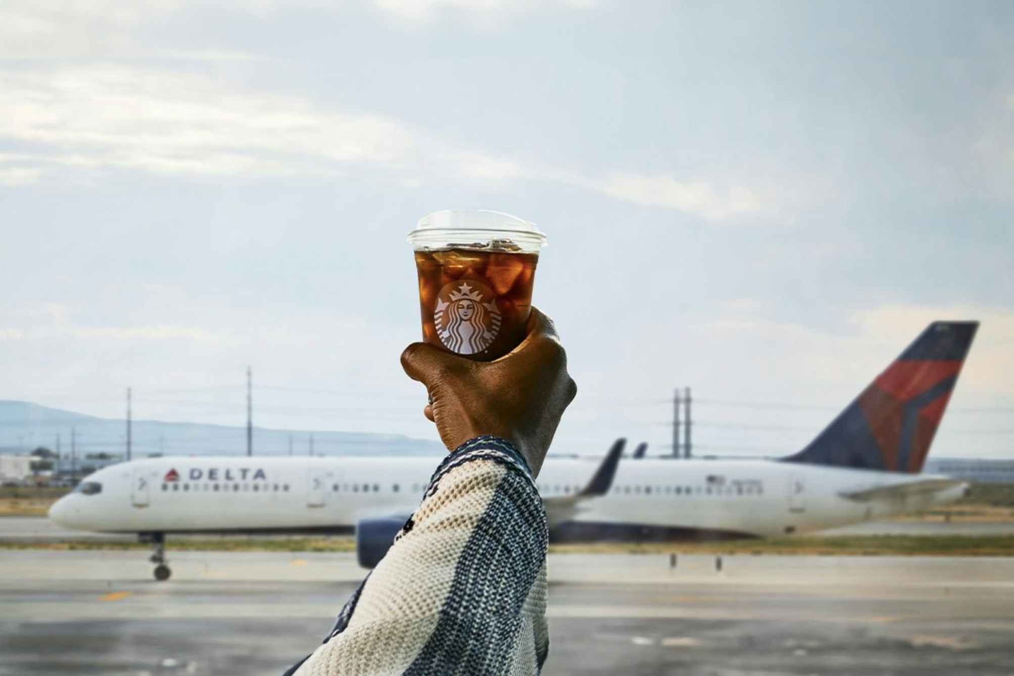 A hand holding a Starbucks coffee cup in front of a Delta Airlines plane