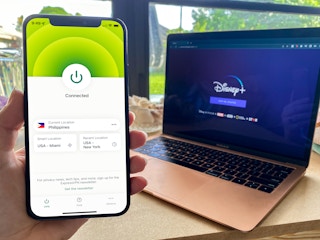 A person holding up a cell phone displaying a connected VPN with a laptop displaying DisneyPlus.com in the background.