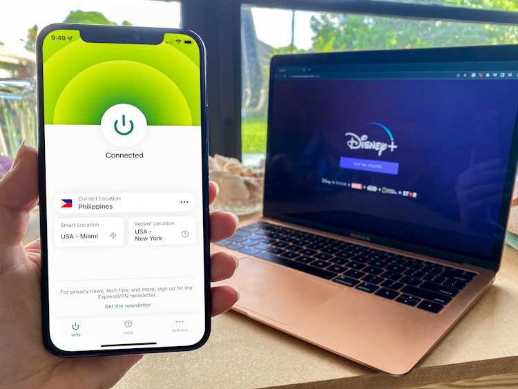 A person holding up a cell phone displaying a connected VPN with a laptop displaying DisneyPlus.com in the background.
