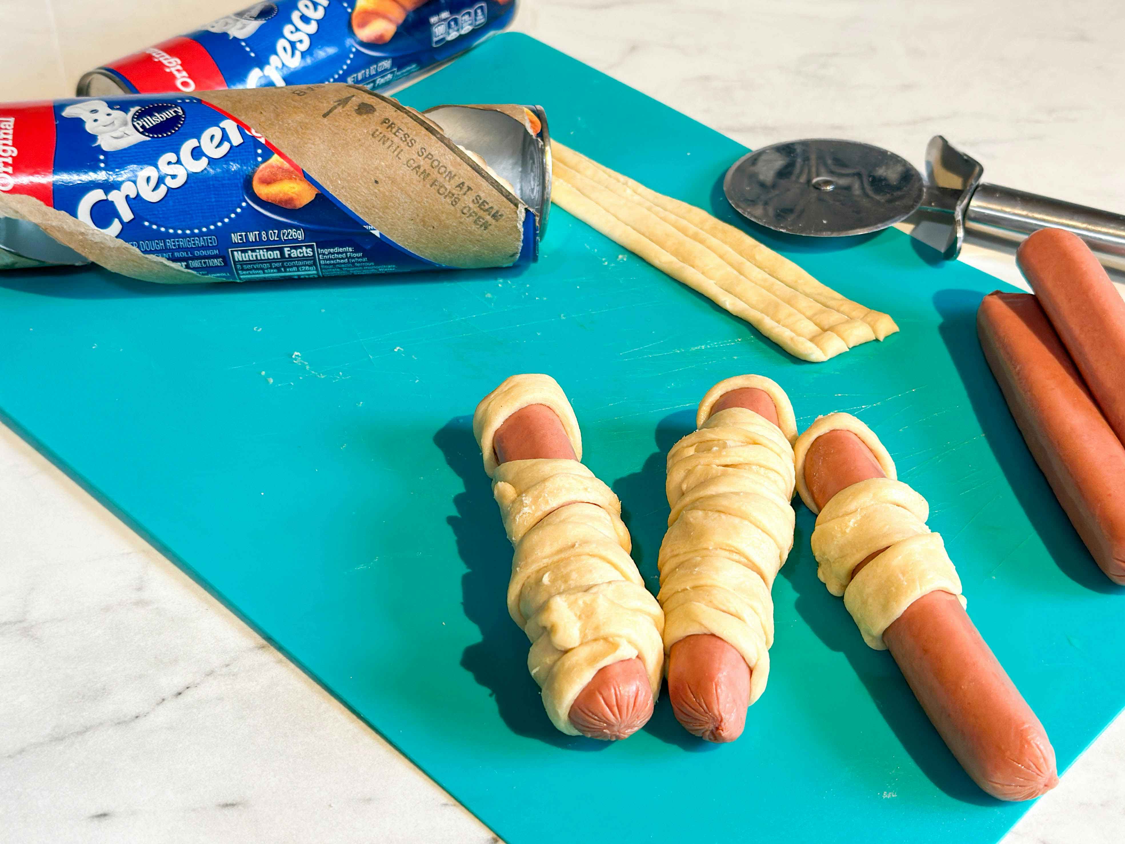 crescent roll dough and hot dogs on a cutting board ready to be baked into hot dog mummy snacks 