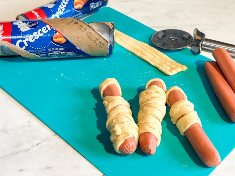 crescent roll dough and hot dogs on a cutting board ready to be baked into hot dog mummy snacks 
