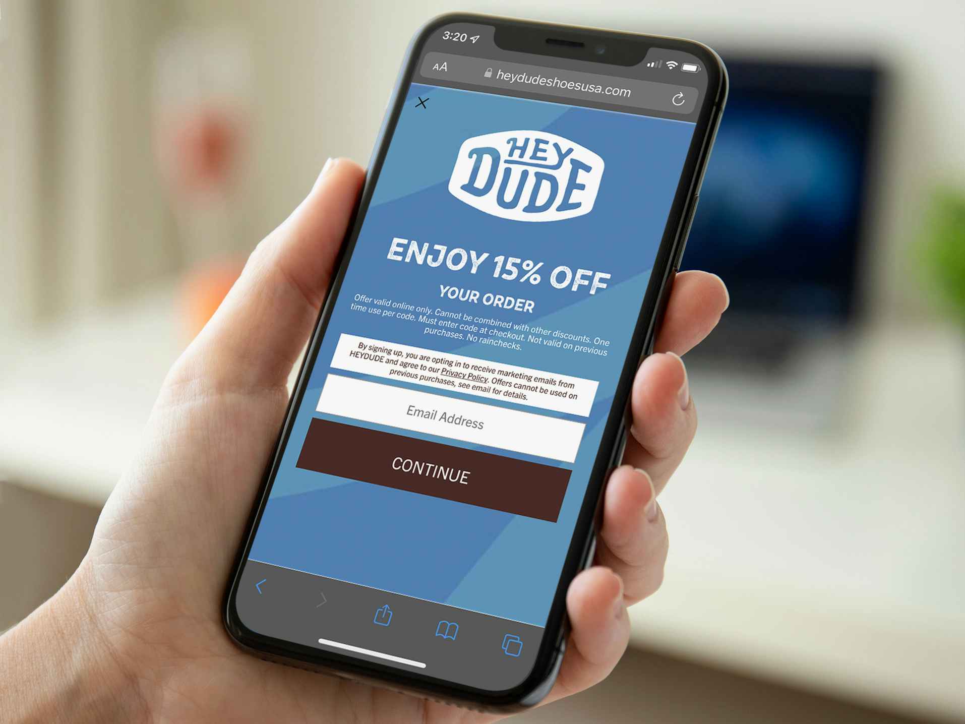 A person's hand holding a phone displaying a coupon on the Hey Dude Shoes USA website for 15% off an order.