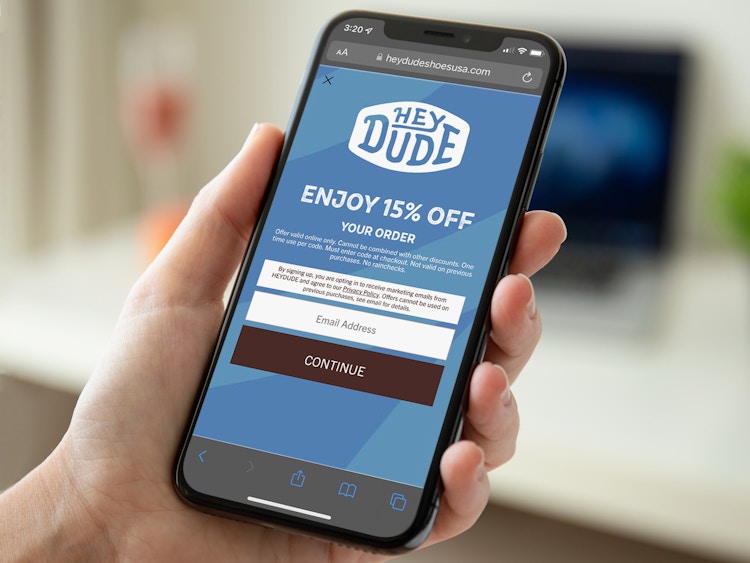 A person's hand holding a phone displaying a coupon on the Hey Dude Shoes USA website for 15% off an order.