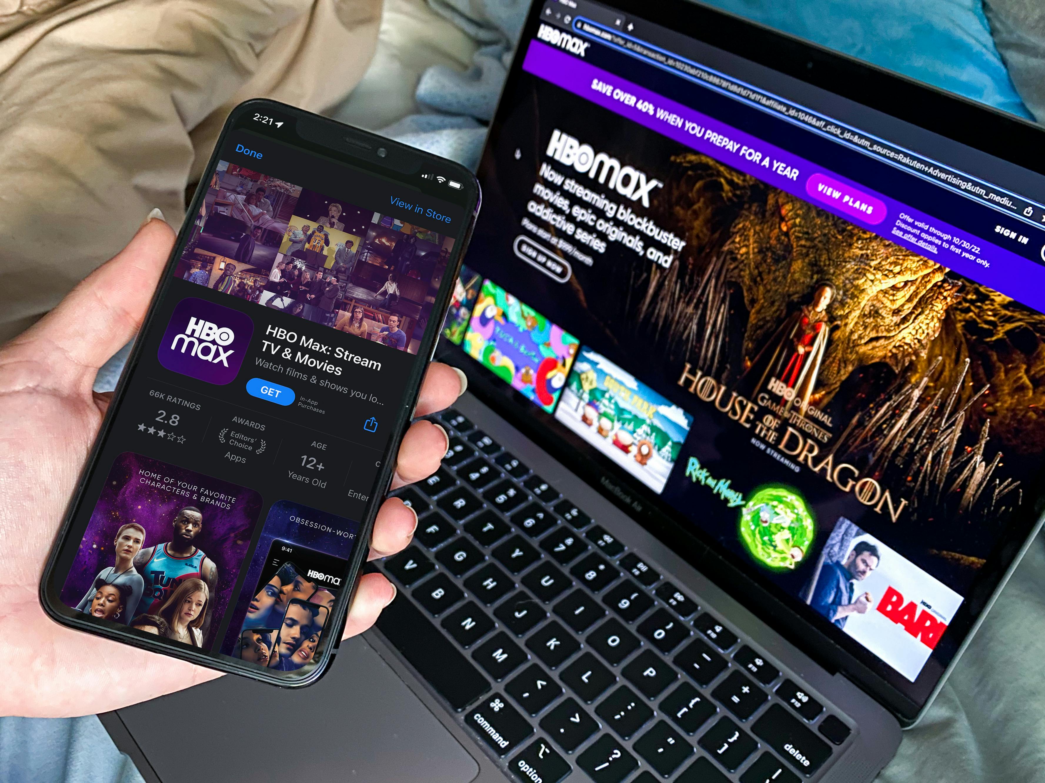 A person's hand holding a phone displaying the HBO Max app download screen near a laptop displaying the banner for House of the Dragon on HBO's website.