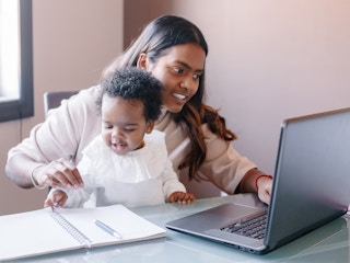 A mom holding her baby and using a laptop on a desk.
