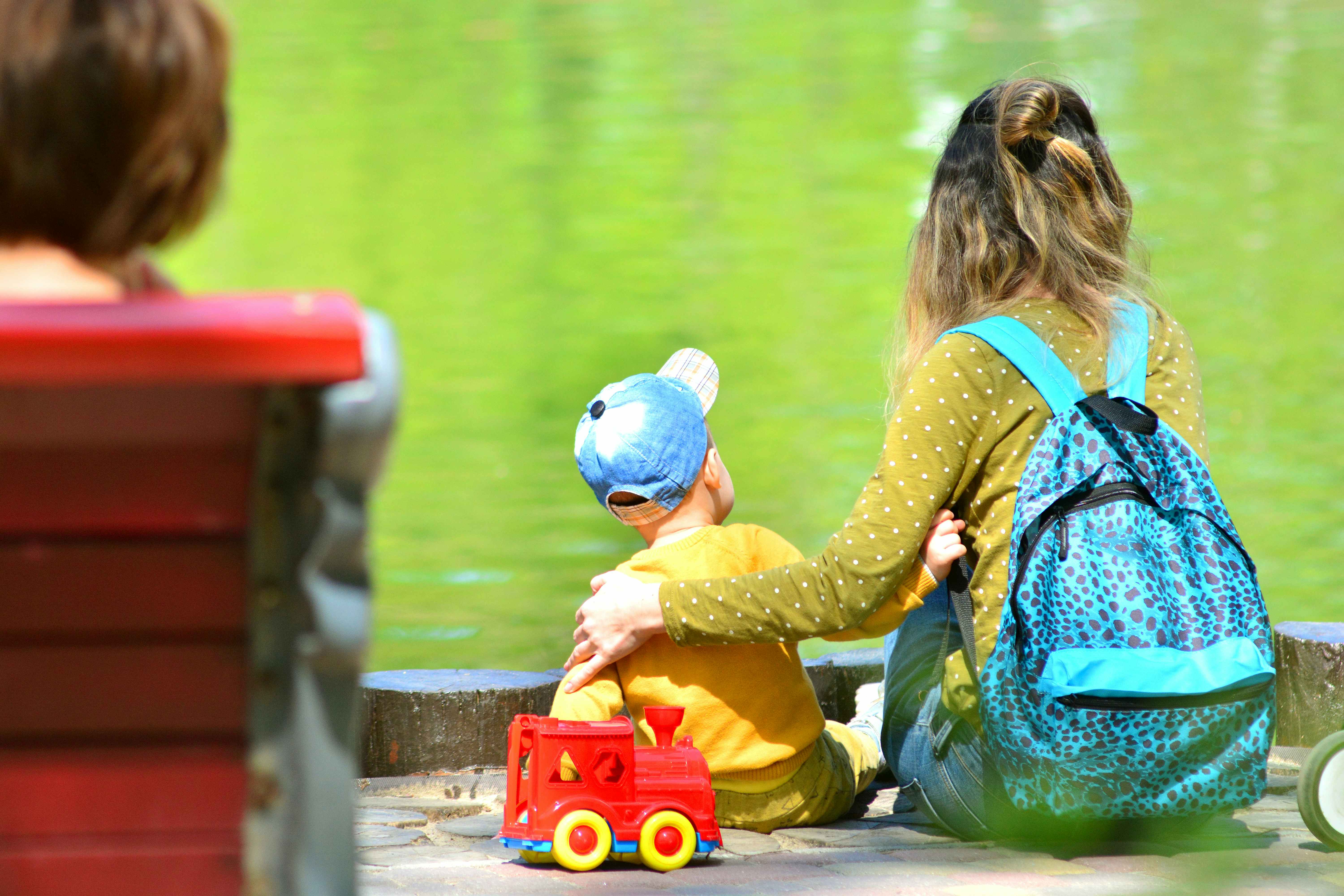 A mom and her child sitting on a curb near some water, the mom wearing a backpack.