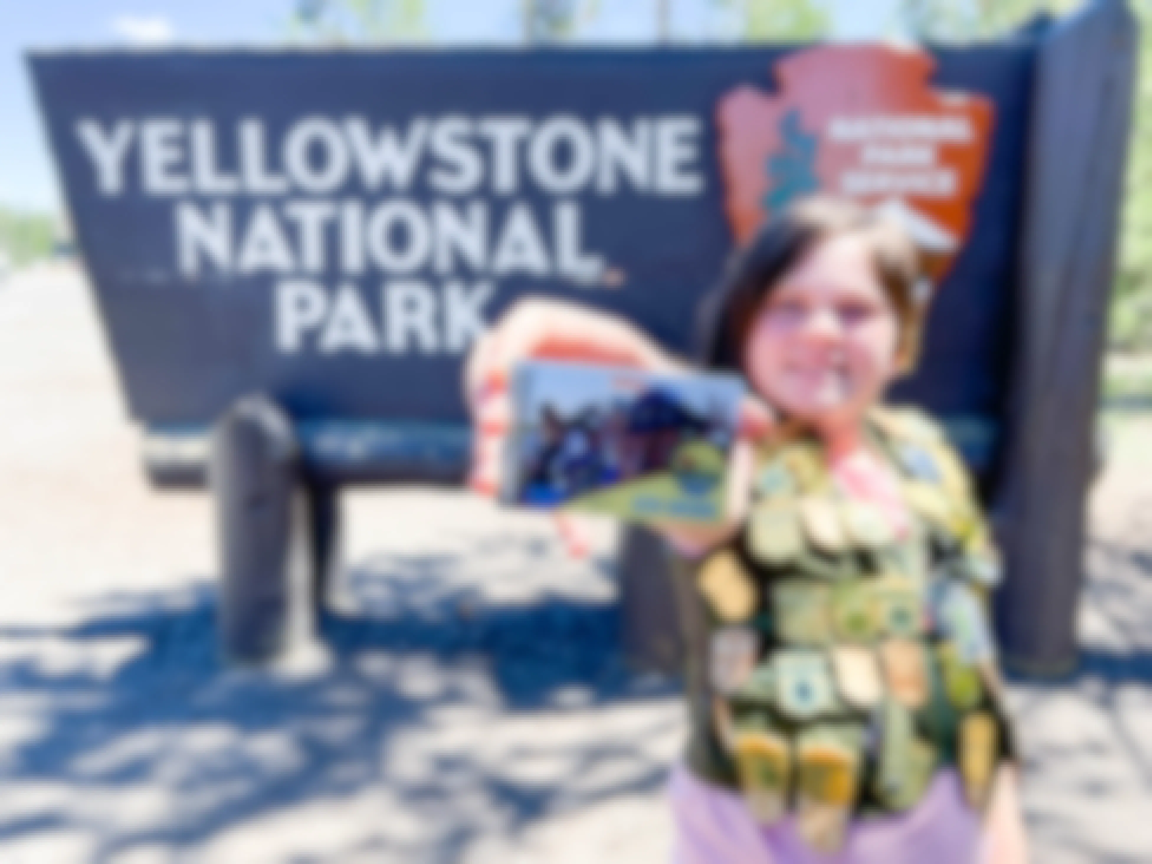A child in front of the yellow stone national park sign wearing a vest covered in junior ranger badges. The child is holding out a free every kids outdoors national park pass.