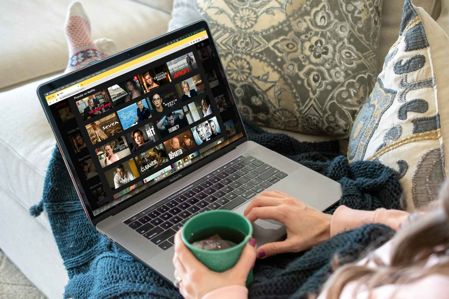 a person holding a cup of tea, laying on a couch, browsing movies on Netflix on a laptop computer.