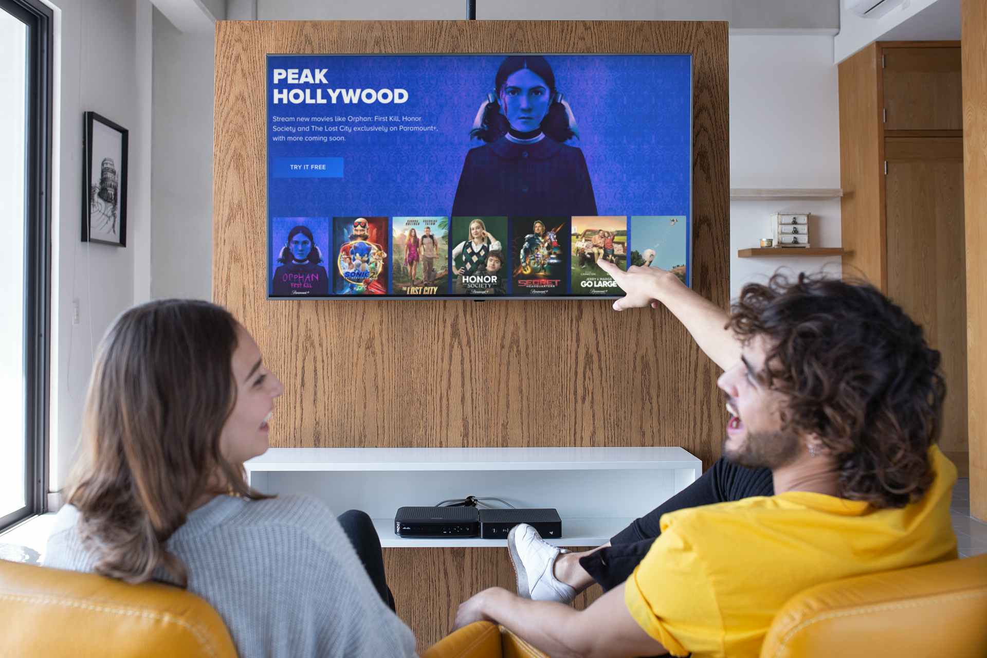 Two people looking at a mounted television. One person wearing a yellow shirt is smiling pointing at the tv screen