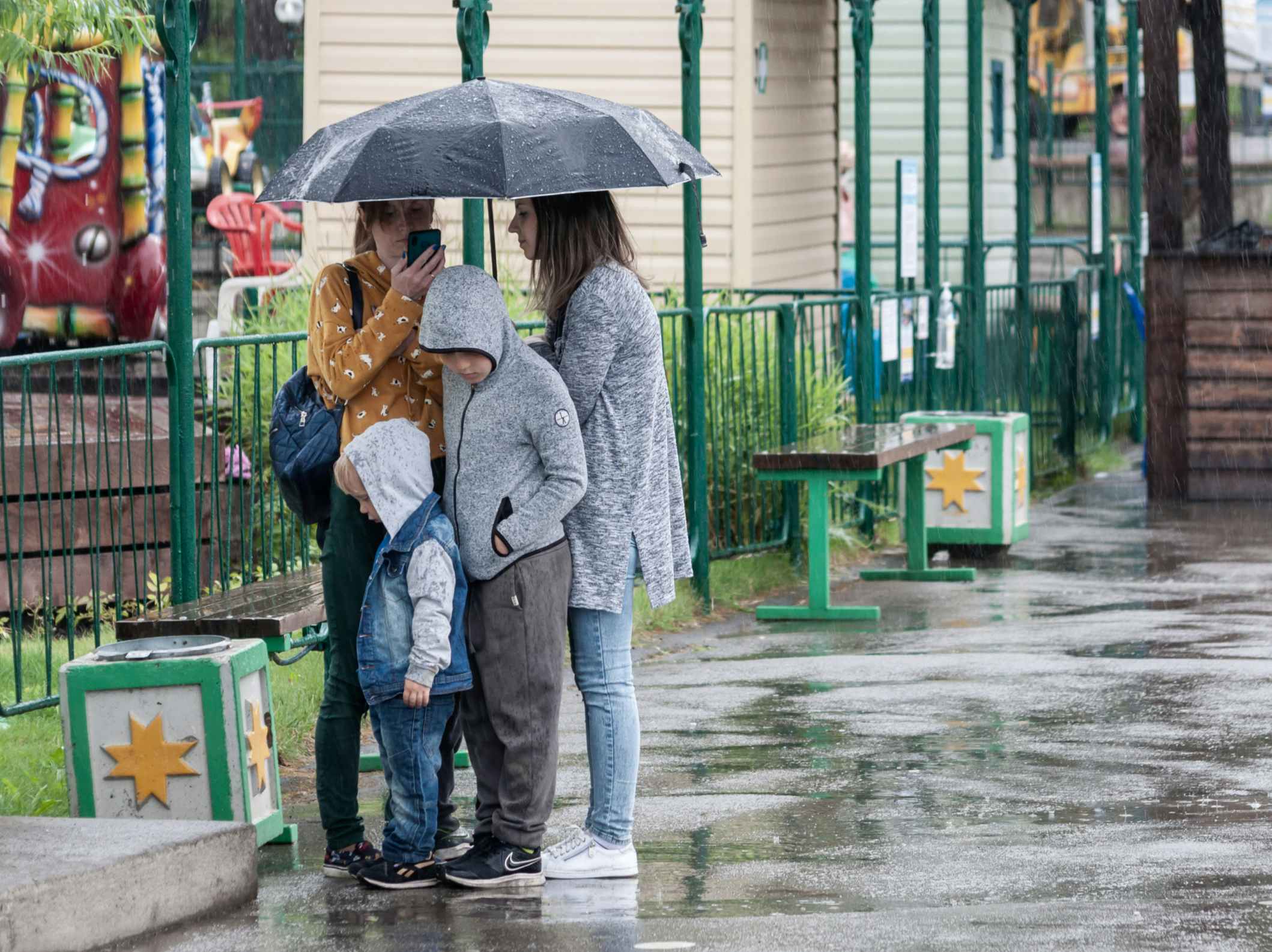 A family grouped under an umbrella on a rainy day at an amusement park.