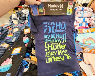 hand holding a 2 pack of kids hurley shirts, front one is navy blue with hurley written numerous times in yellow and blue