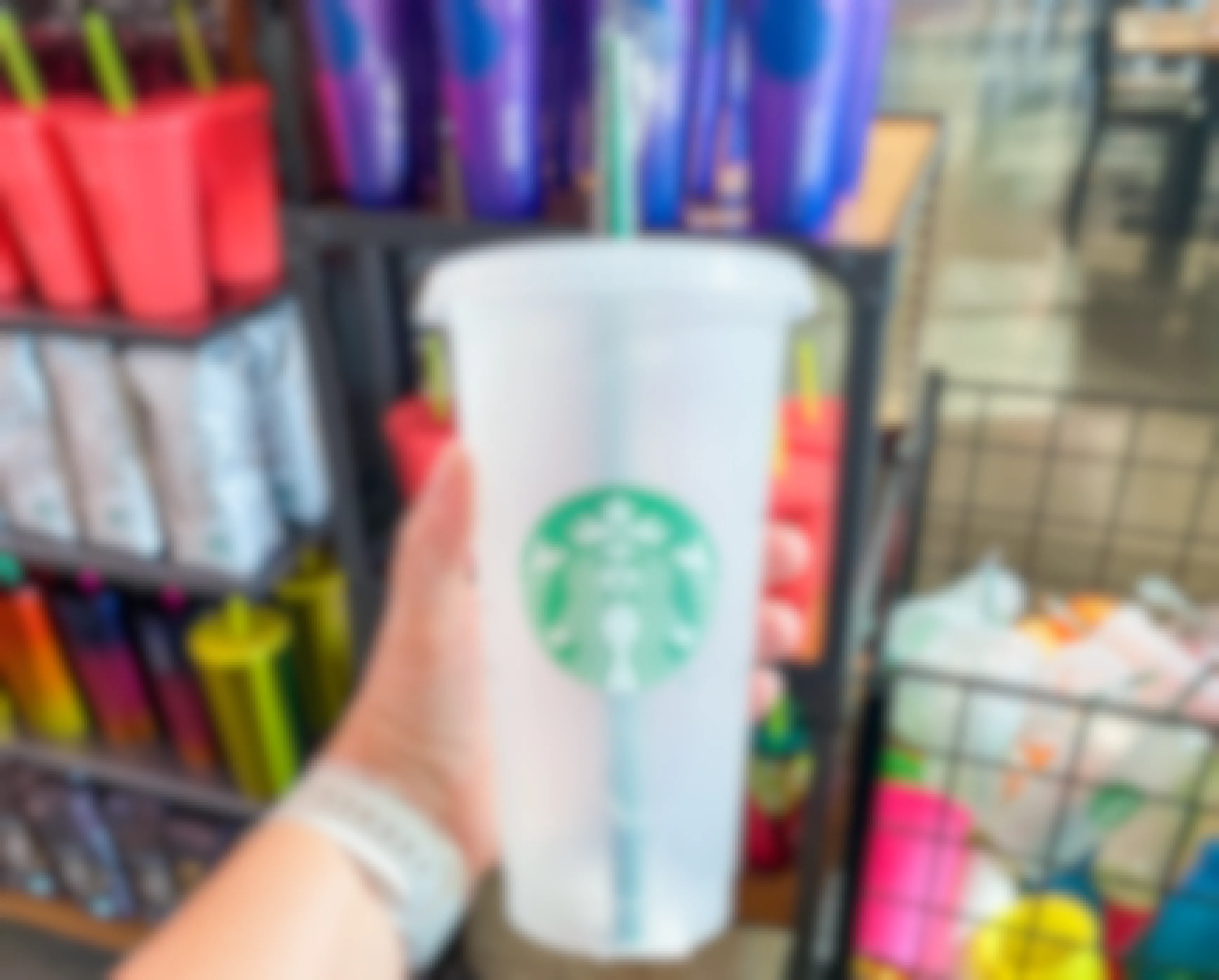 A person's hand holding up a reuasble Starbucks tumbler cup in front of a shelf of more cups.