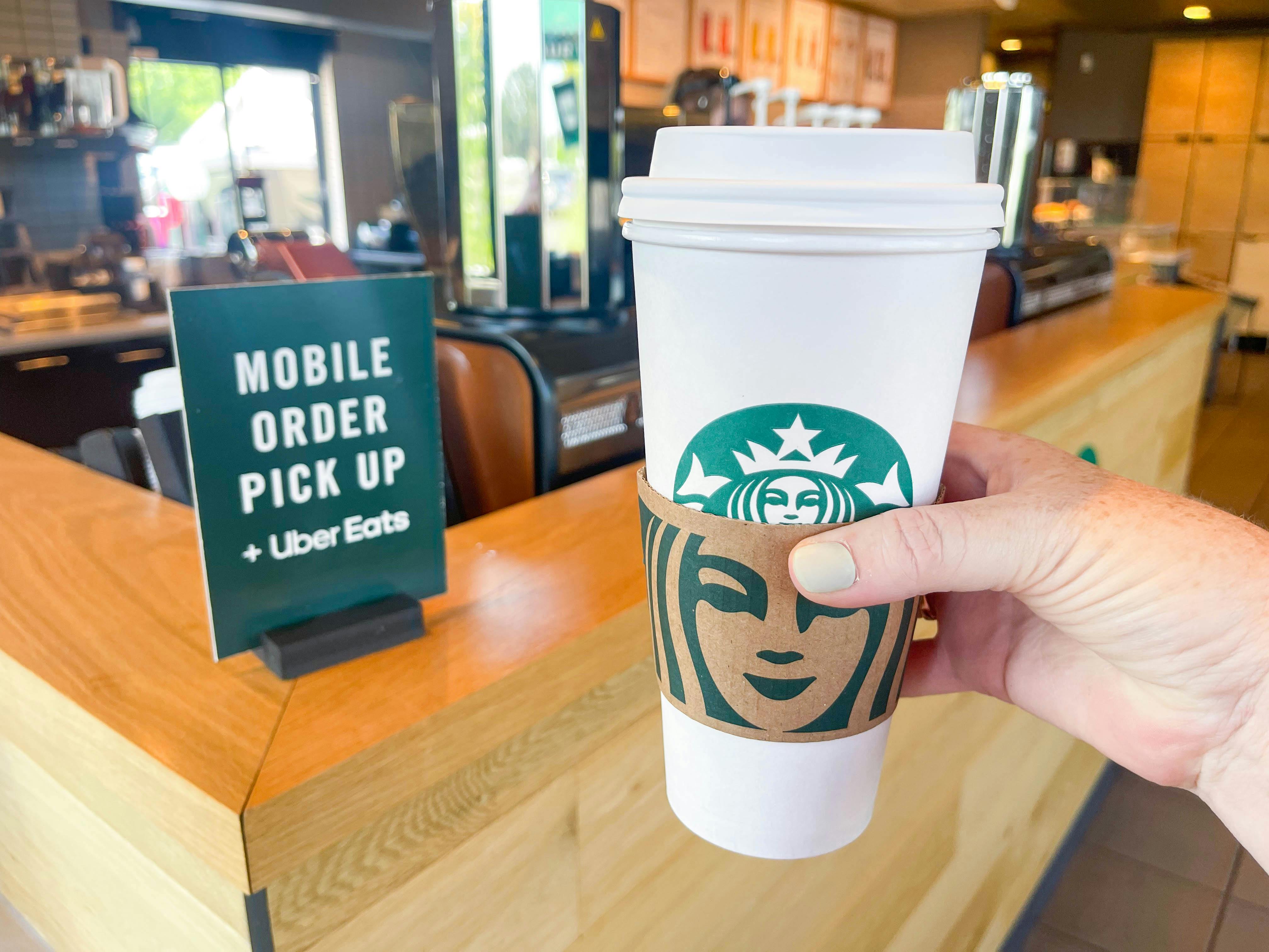A person holding a large Starbucks hot beverage near the Mobile Order Pick Up sign inside Starbucks