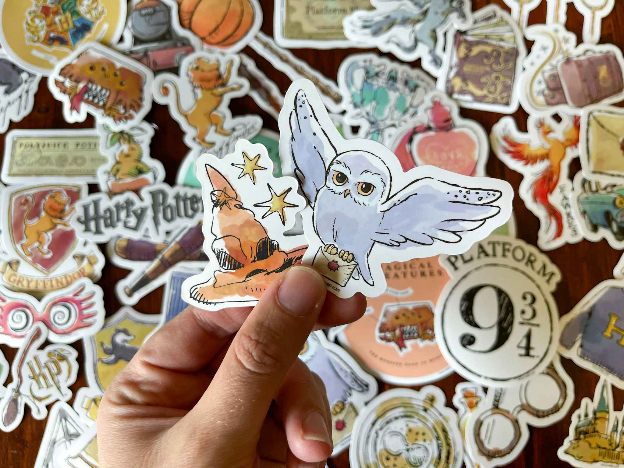 A person holding two Harry Potter themed vinyl stickers with dozens of others laying out on a table in the background.