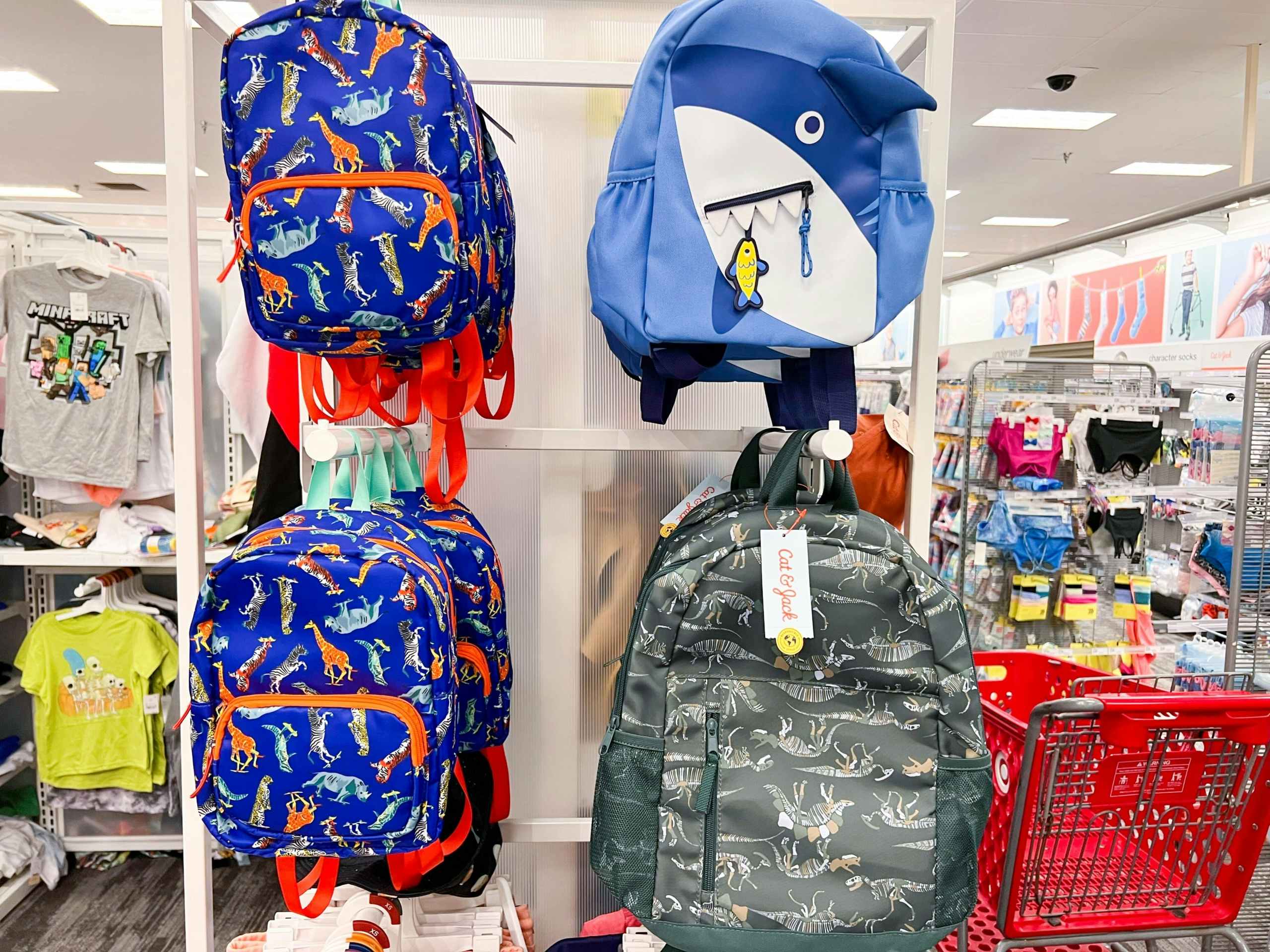 a blue shark, blue dinosaur, and green dinosaur backpacks hanging on a wall at Target