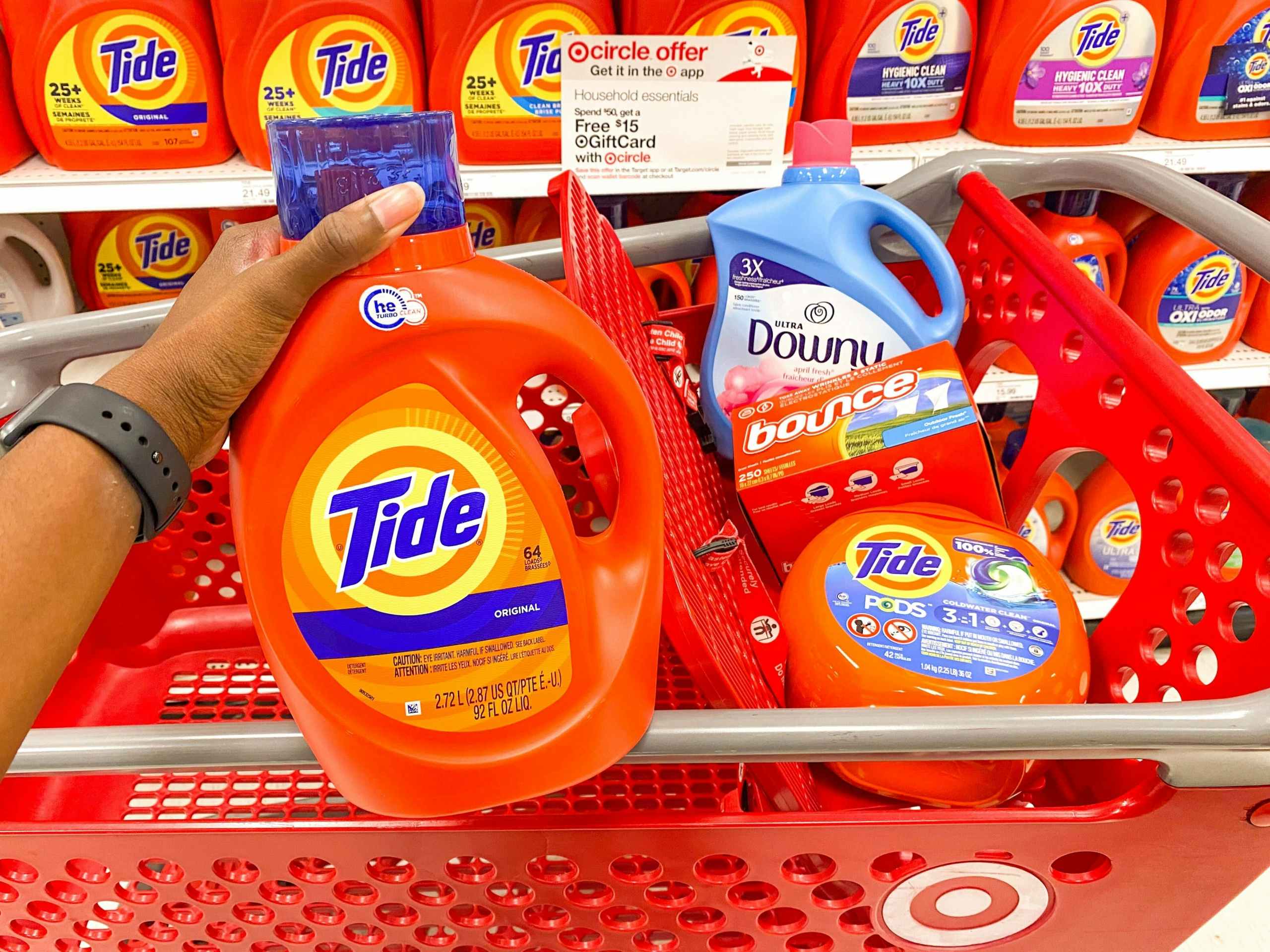 hand holding up a bottle of Tide in a Target aisle in front of a shopping cart that contains Tide pods, Downy liquid, and Bounce dryer sheets
