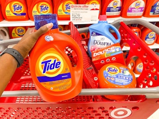 hand holding up a bottle of Tide in a Target aisle in front of a shopping cart that contains Tide pods, Downy liquid, and Bounce dryer sheets