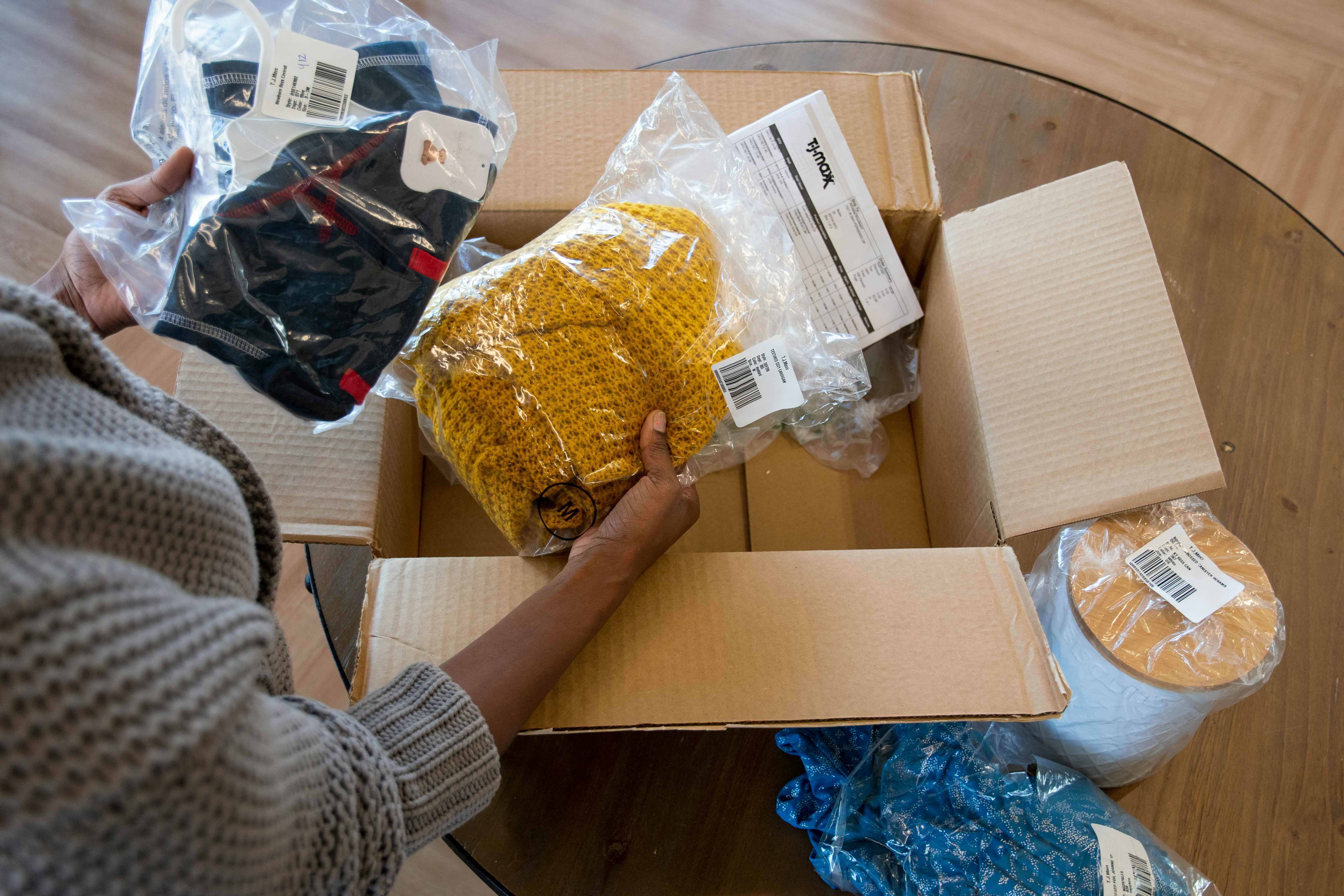A woman pulling products wrapped in plastic from a large cardboard box. A T.J.Maxx online order receipt is visible in the background.
