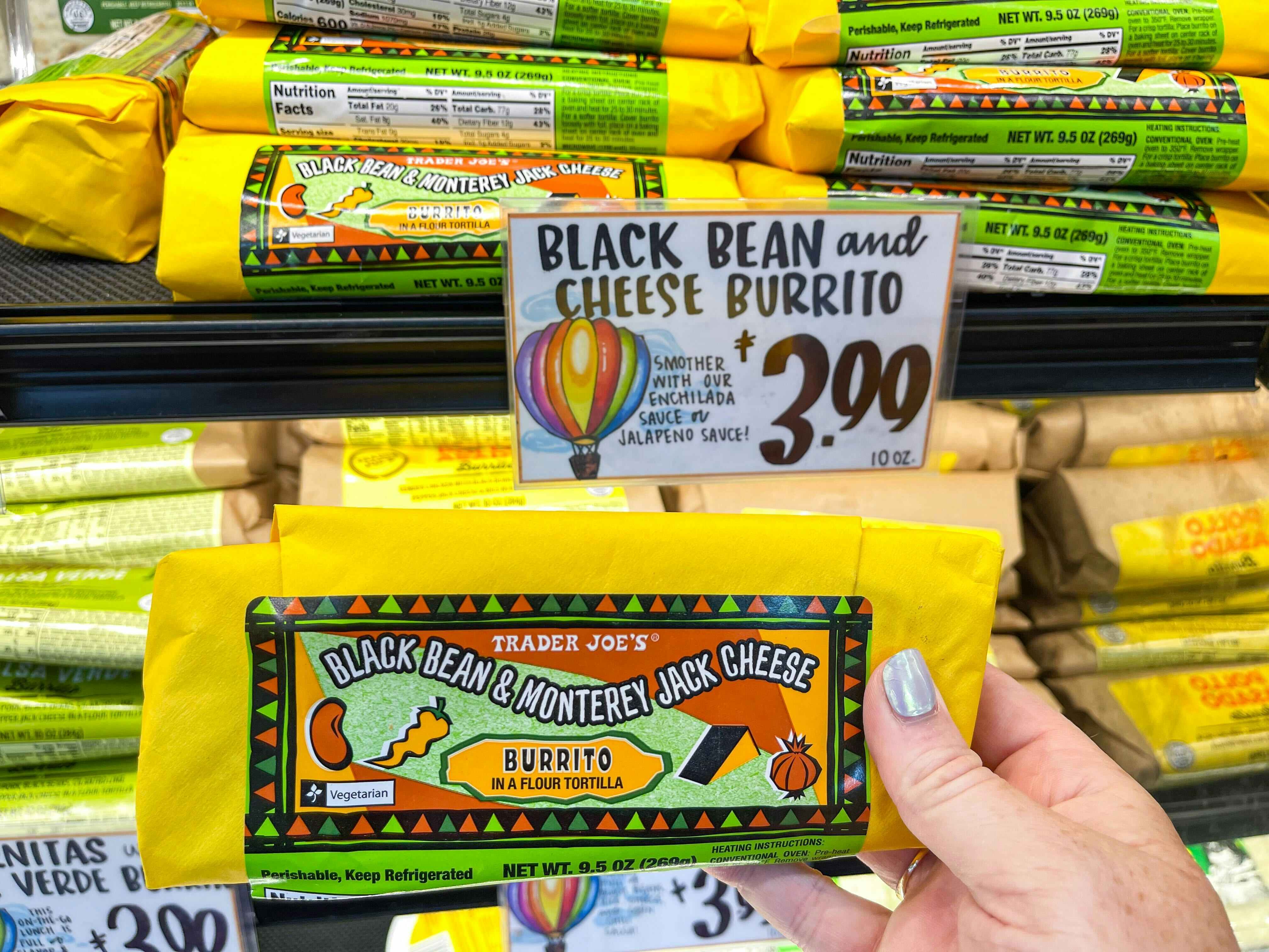 A person's hand holding a Trader Joe's Black Bean and Cheese Burrito in front of a shelf of them inside Trader Joe's.