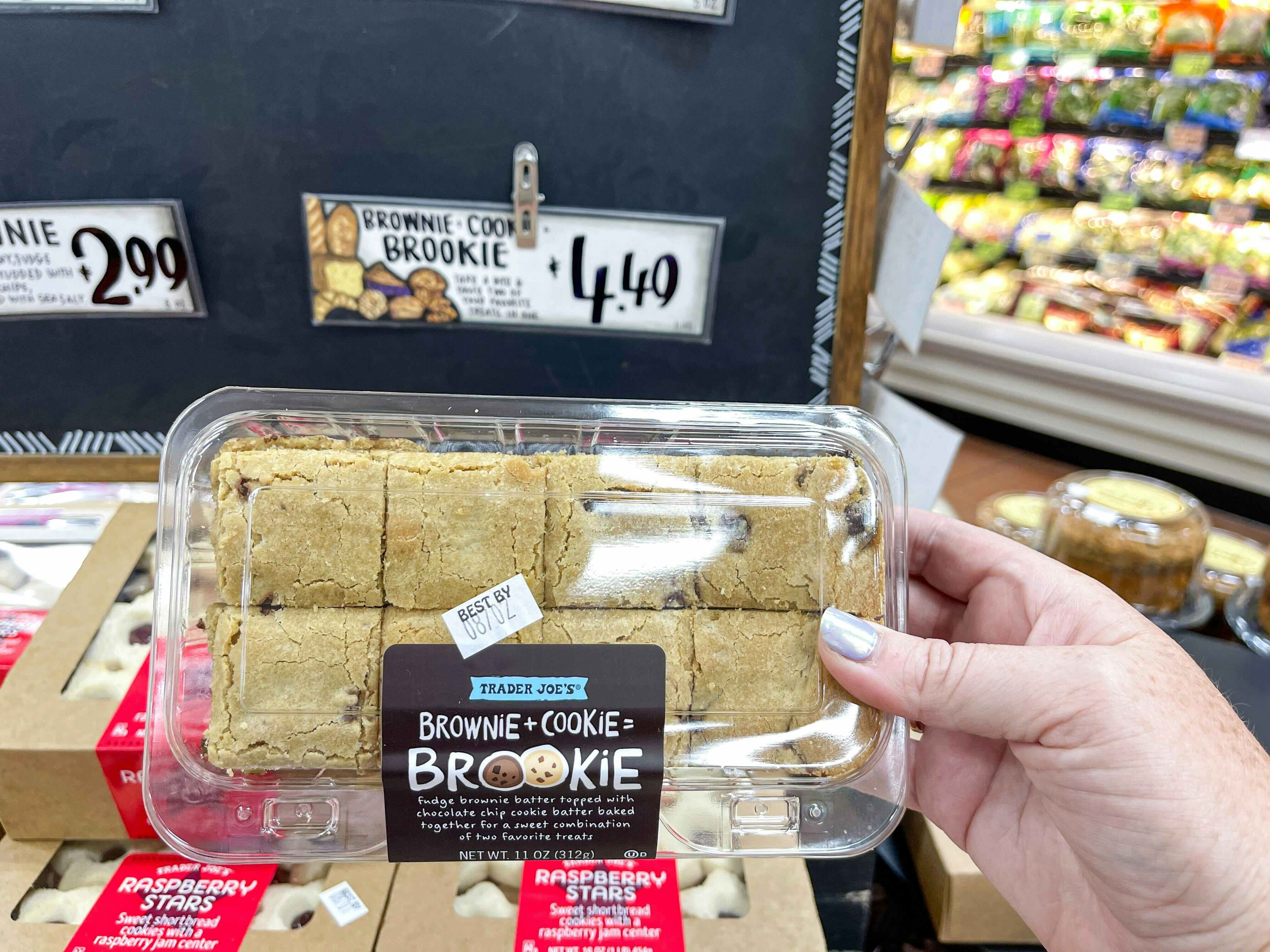 A person's hand holding a container of Trader Joe's Brookie in front of a table inside Trader Joe's.