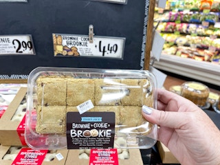 A person's hand holding a container of Trader Joe's Brookie in front of a table inside Trader Joe's.