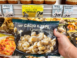 A person's hand holding a bag of Trader Joe's Cacio E Pepe Gnocchi above a display of them inside Trader Joe's.