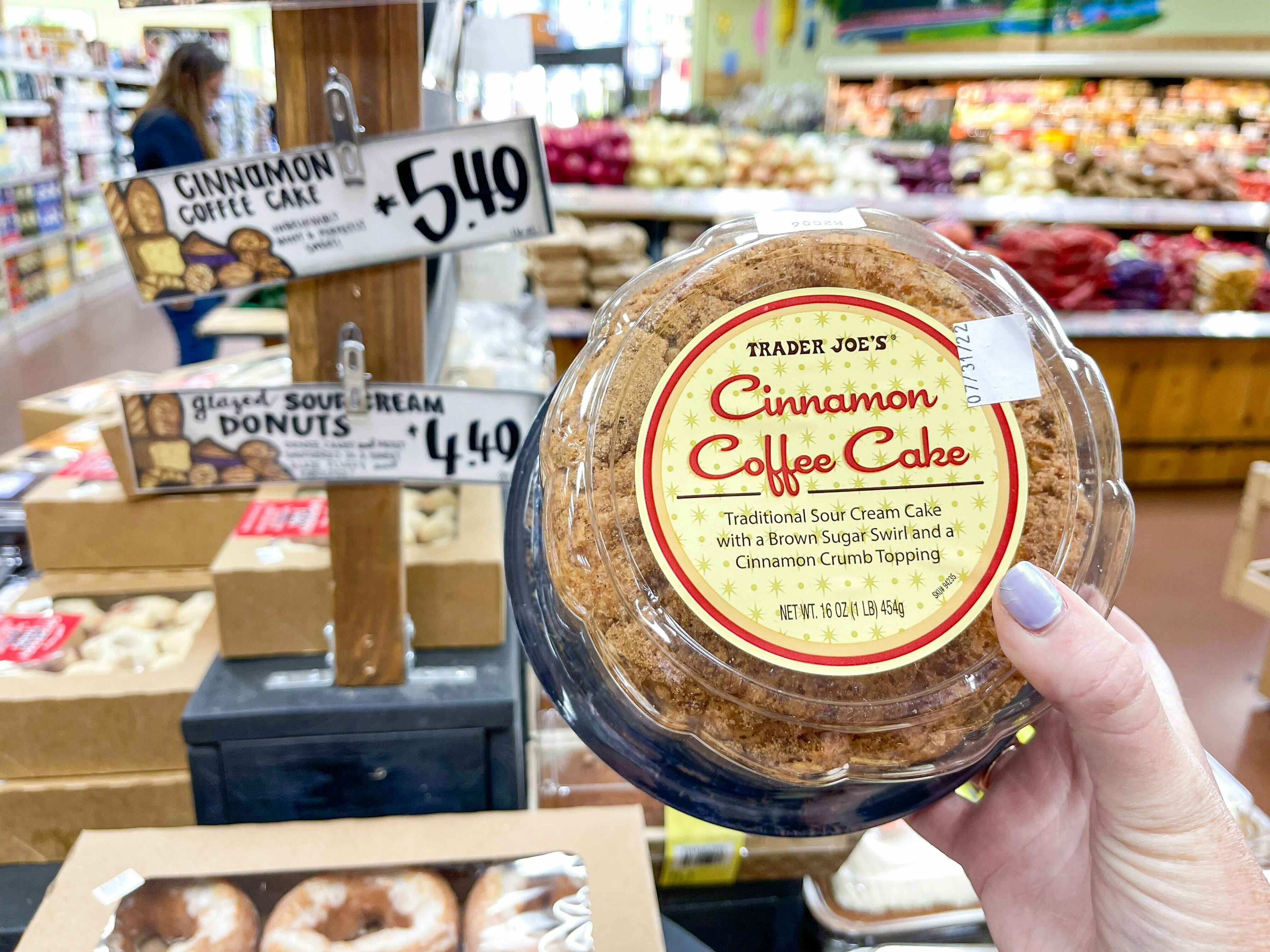A person's hand holding a Trader Joe's Cinnamon Coffee Cake in front of a table in the bakery at Trader Joe's.