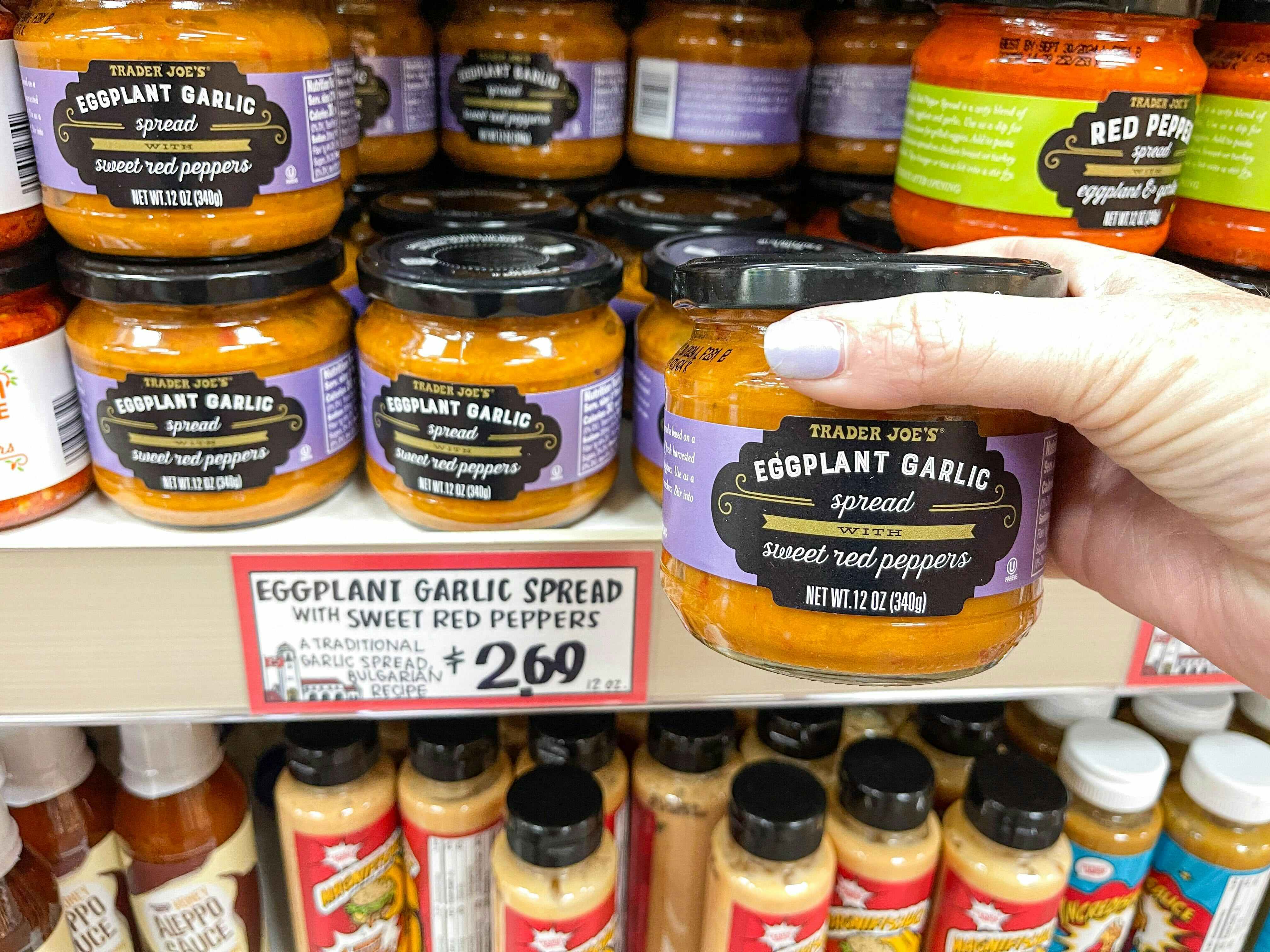 A person's hand holding a jar of Trader Joe's Eggplant Garlic Spread with Sweet Red Peppers in front of a shelf of them at Trader Joe's.