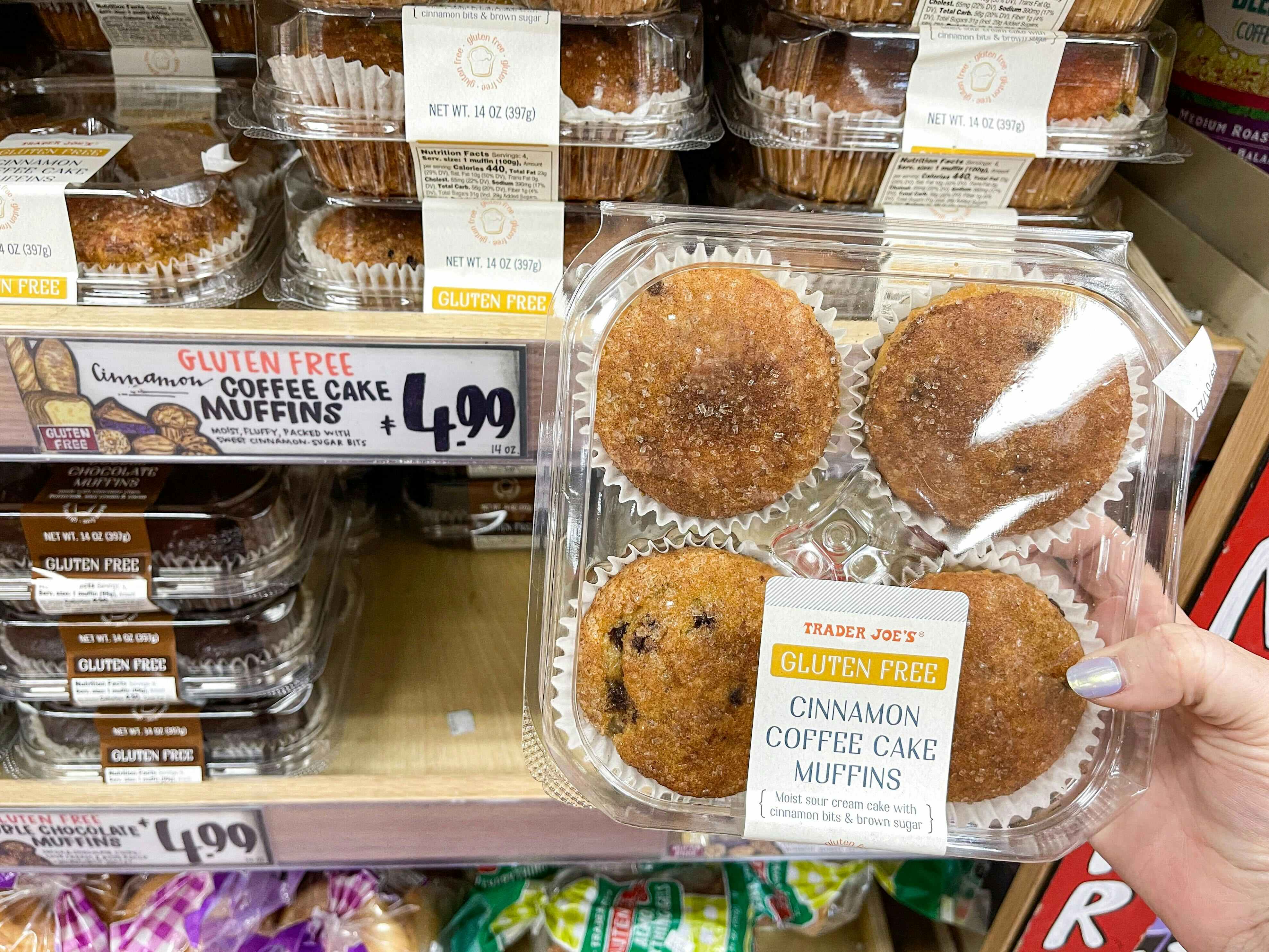 A person's hand holding a container of Cinnamon Coffee Cake Muffins in front of a shelf of them at Trader Joe's.