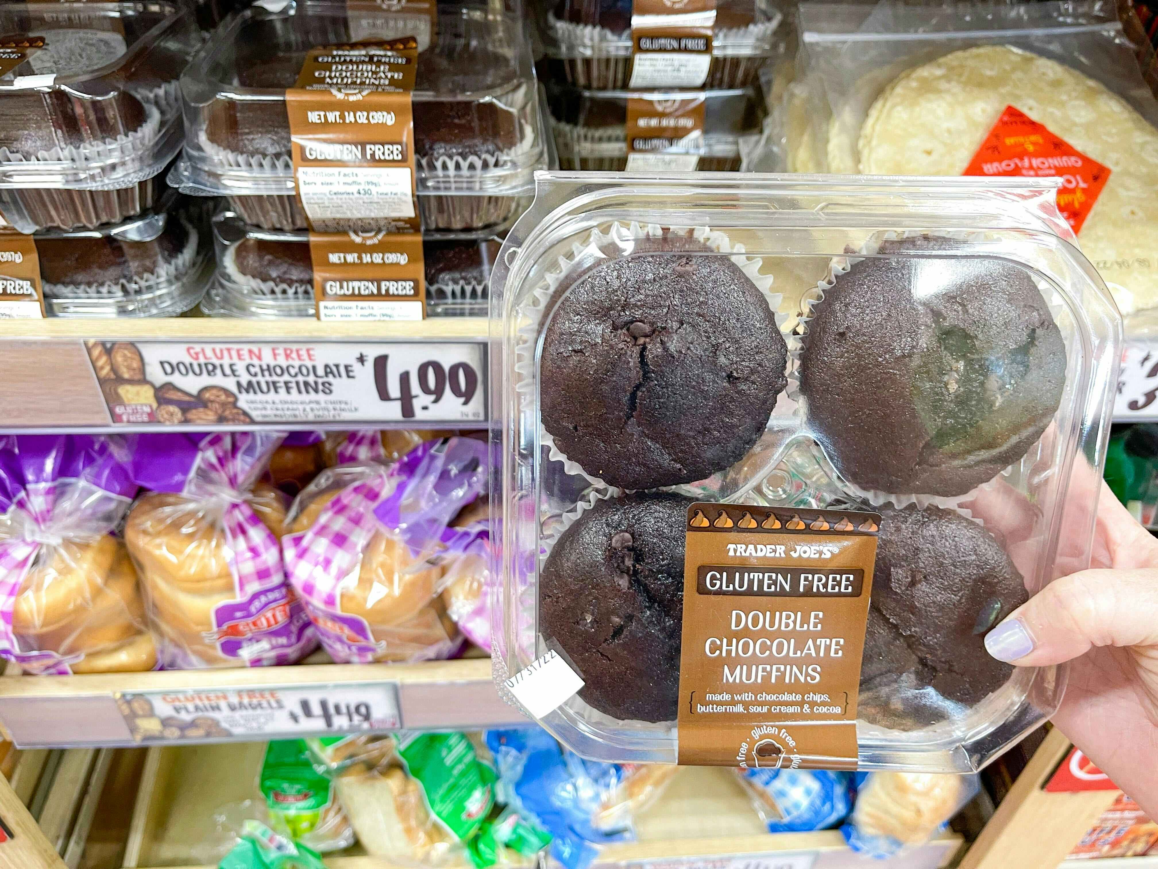 A person's hand holding a container of Trader Joe's Gluten Free Double Chocolate Muffins in front of a shelf of them at Trader Joe's.