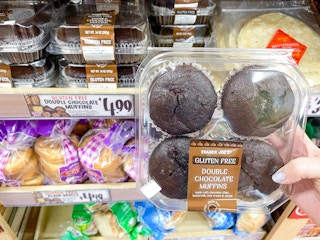 A person's hand holding a container of Trader Joe's Gluten Free Double Chocolate Muffins in front of a shelf of them at Trader Joe's.