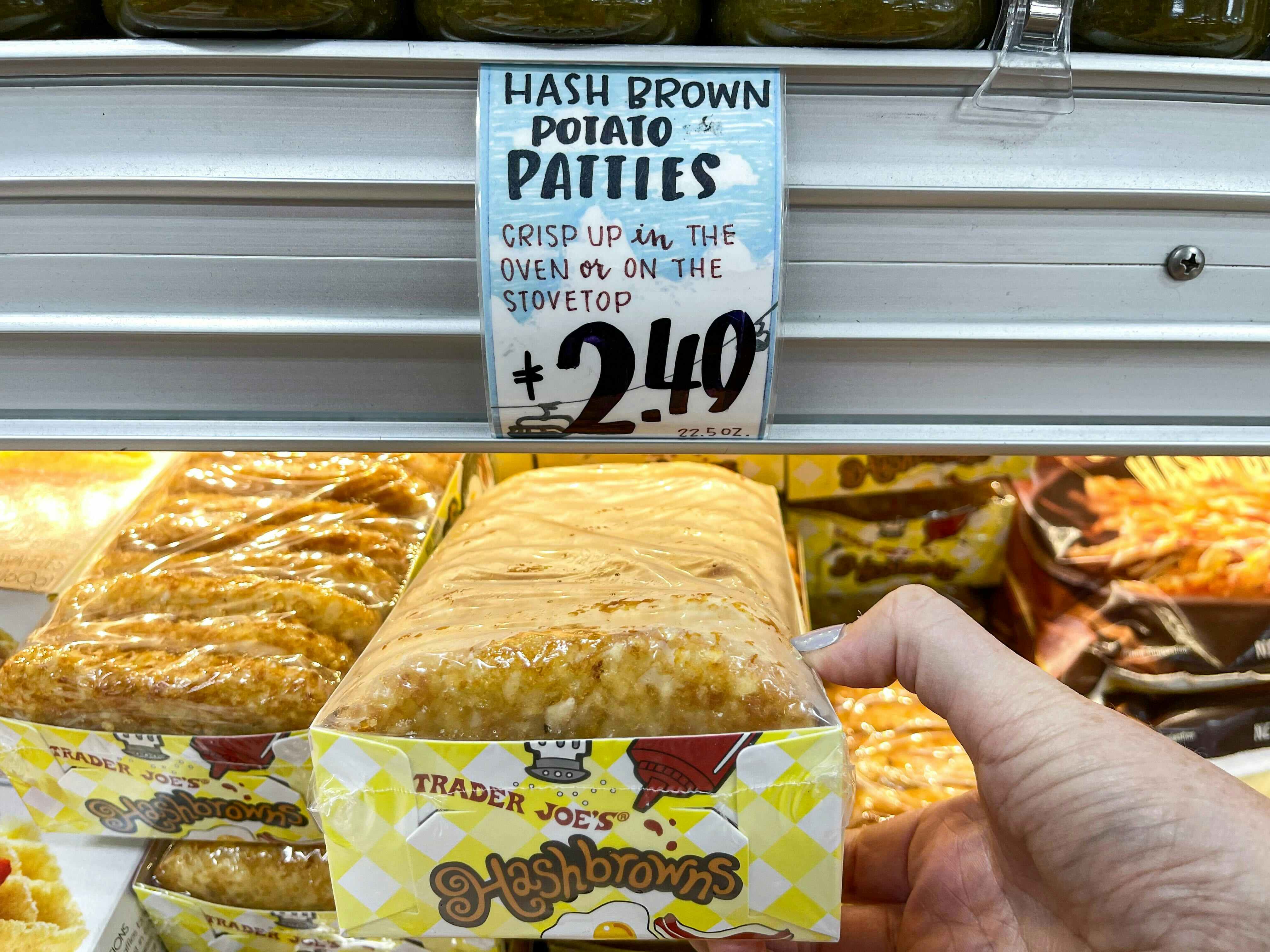 A person's hand holding a package of Trader Joe's Hashbrowns in front of the price sign on the shelf.