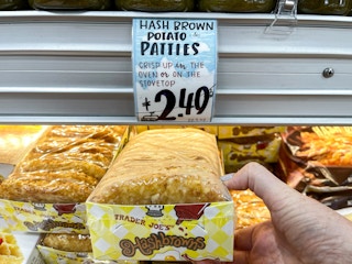 A person's hand holding a package of Trader Joe's Hashbrowns in front of the price sign on the shelf.